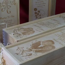 Drewniane pudełka z grawerowanymi dedykacjami, zdjęciami i ornamentami, ustawione obok butelki wina i dekoracyjnego wypełniacza.
