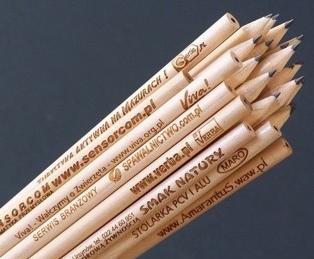 Stos drewnianych ołówków z nadrukowanymi adresami stron internetowych i logotypami firm, ułożonych pod kątem na ciemnym tle.