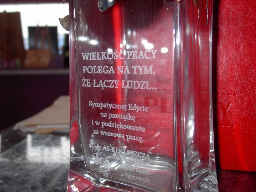 Szklana statuetka z wygrawerowanym tekstem dedykacji i podziękowaniami za wzorową pracę, umieszczona na blacie.