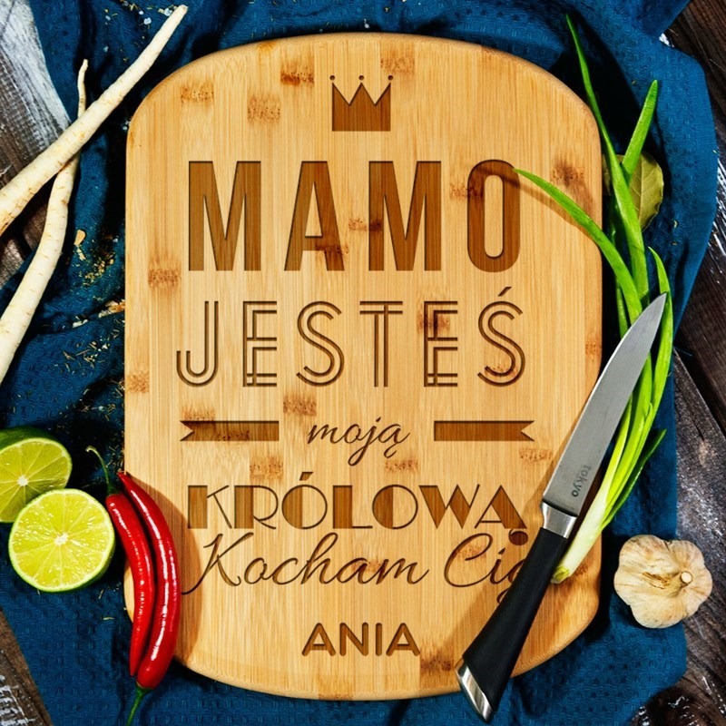 Deska bambusowa z wygrawerowanym napisem 'Mamo Jesteś moją Królową Kocham Cię Ania' otoczona warzywami i nożem na niebieskiej ściereczce.