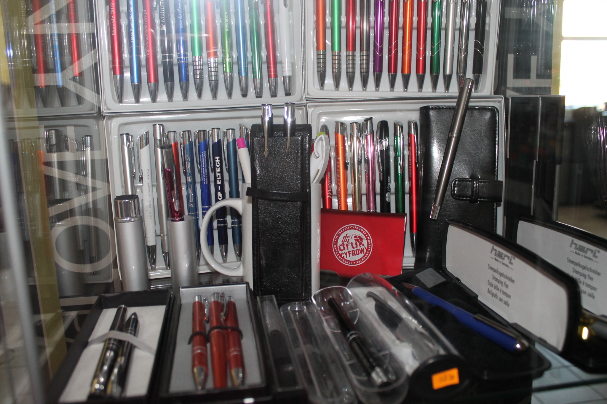 Ekspozycja różnorodnych długopisów reklamowych w pudełkach i etui, w tym metalowe, kolorowe i z nadrukiem, prezentowanych za szybą witryny sklepowej.