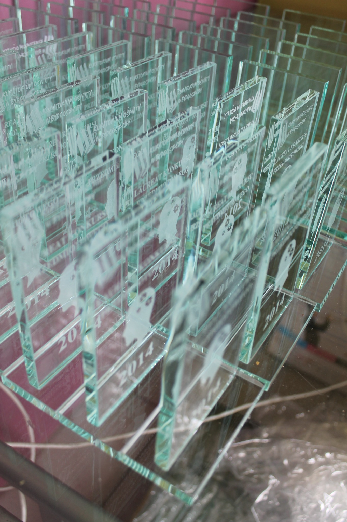 Szereg szklanych statuetek z wygrawerowanymi napisami i grafikami, upamiętniającymi konkurs z 2014 roku, ustawionych na szklanej powierzchni.