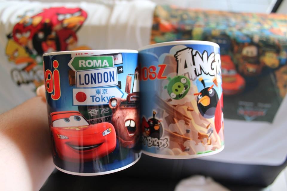 Dwa kubki z nadrukiem: jeden z motywami z filmu Auta (Zygzak McQueen, Złomek i drogowskazy na Rzym, Londyn i Tokio), drugi z postaciami z gry Angry Birds, na rozmytym tle koszulka i plakat z tymi...