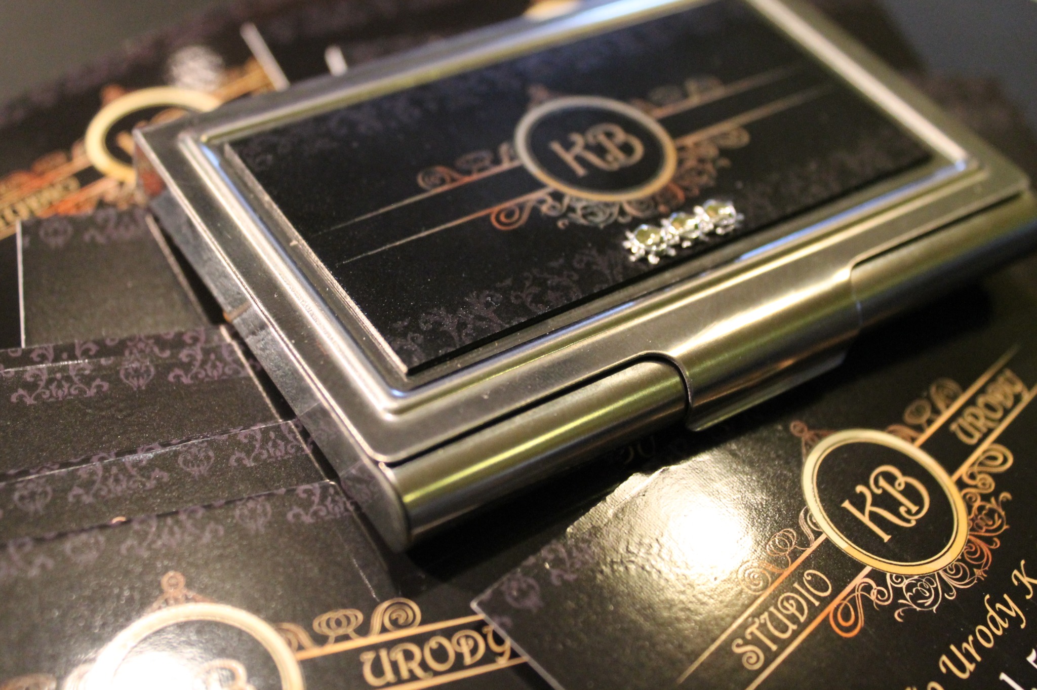 Metalowe etui na karty z dekoracyjnymi kryształkami leży na stercie czarnych kart z logo 'KB Studio Urody' w złotej obwódce i ozdobnymi ornamentami.