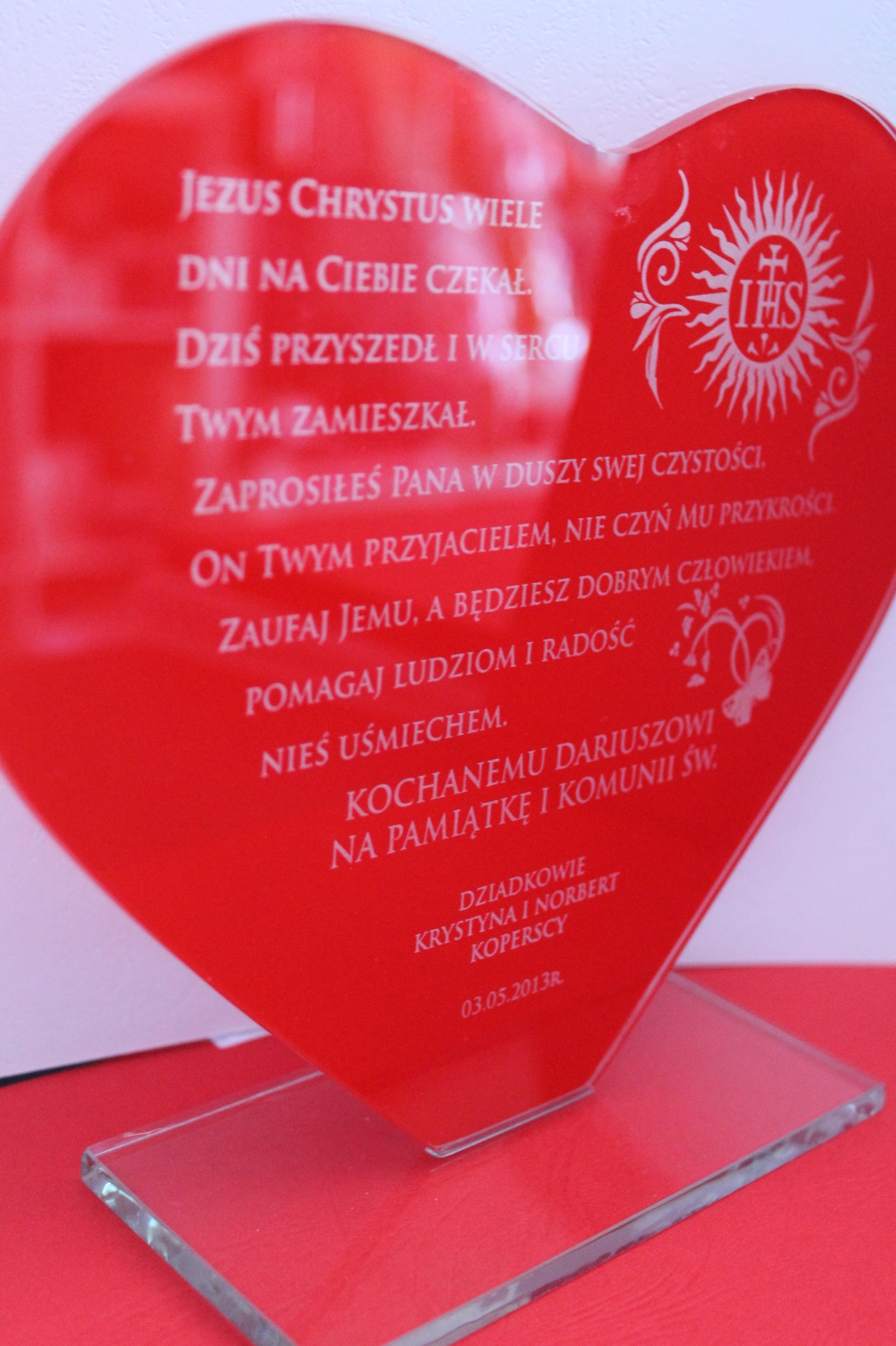 Czerwona statuetka w kształcie serca z białym nadrukiem tekstu religijnego i symbolu IHS, umieszczona na przezroczystej podstawce z pleksi, pamiątka z okazji Pierwszej Komunii Świętej.
