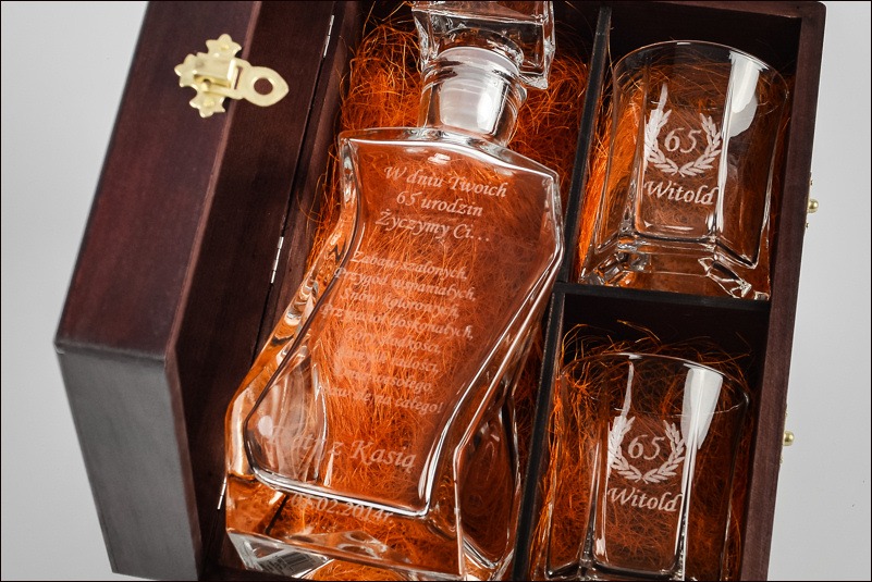 Karafka i dwa szklanki z grawerem dedykacji w drewnianym pudełku zamykanym na ozdobny zatrzask. Na karafce wygrawerowane życzenia urodzinowe, a na szklankach imię 'Witold' i liczba 65 w wieńcu...