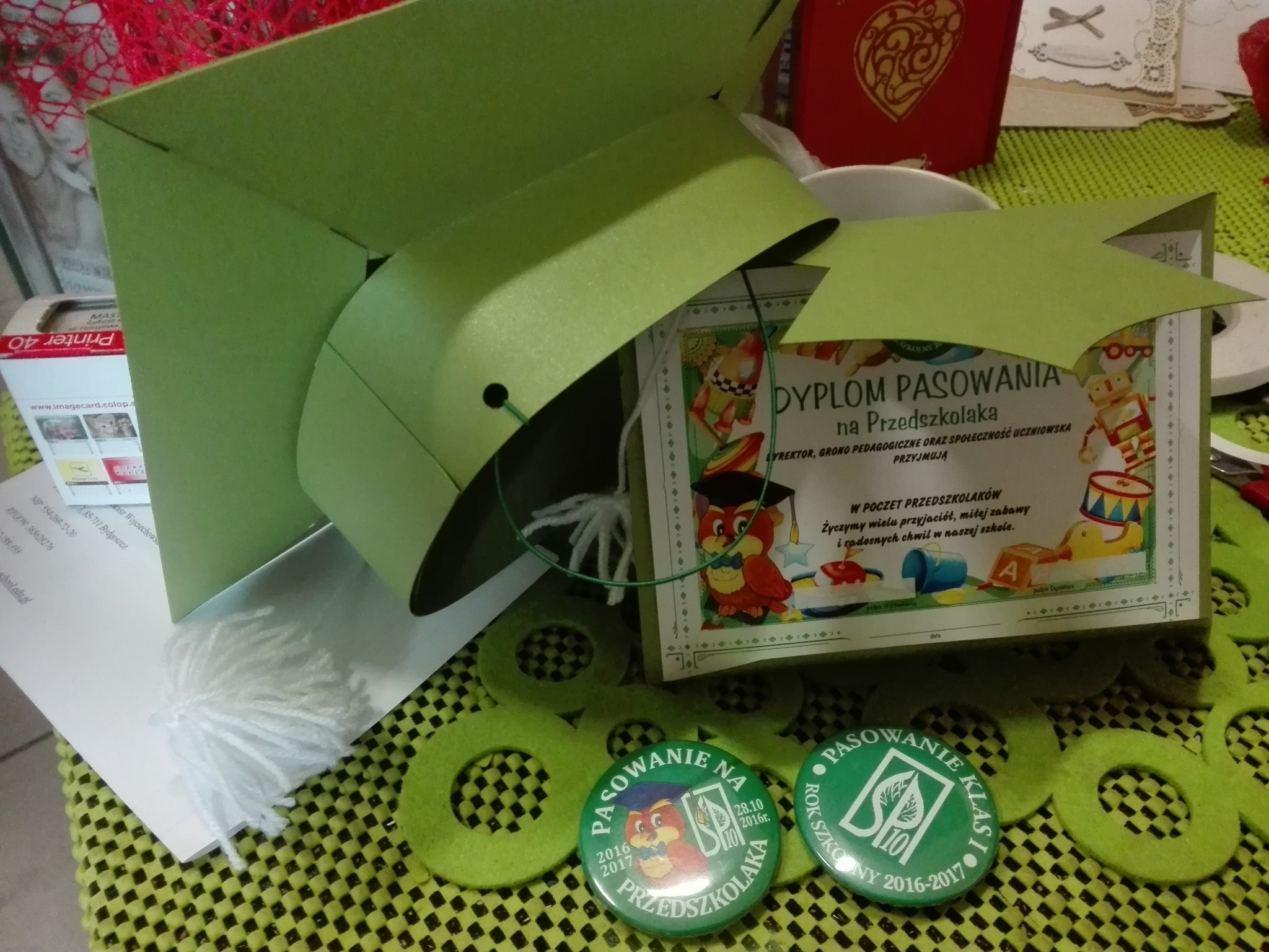 Ręcznie robione elementy dekoracyjne i pamiątkowe z okazji pasowania na przedszkolaka: dyplom, czapka absolwenta, znaczki i ozdobne kółka z filcu, prezentowane na zielonej macie.