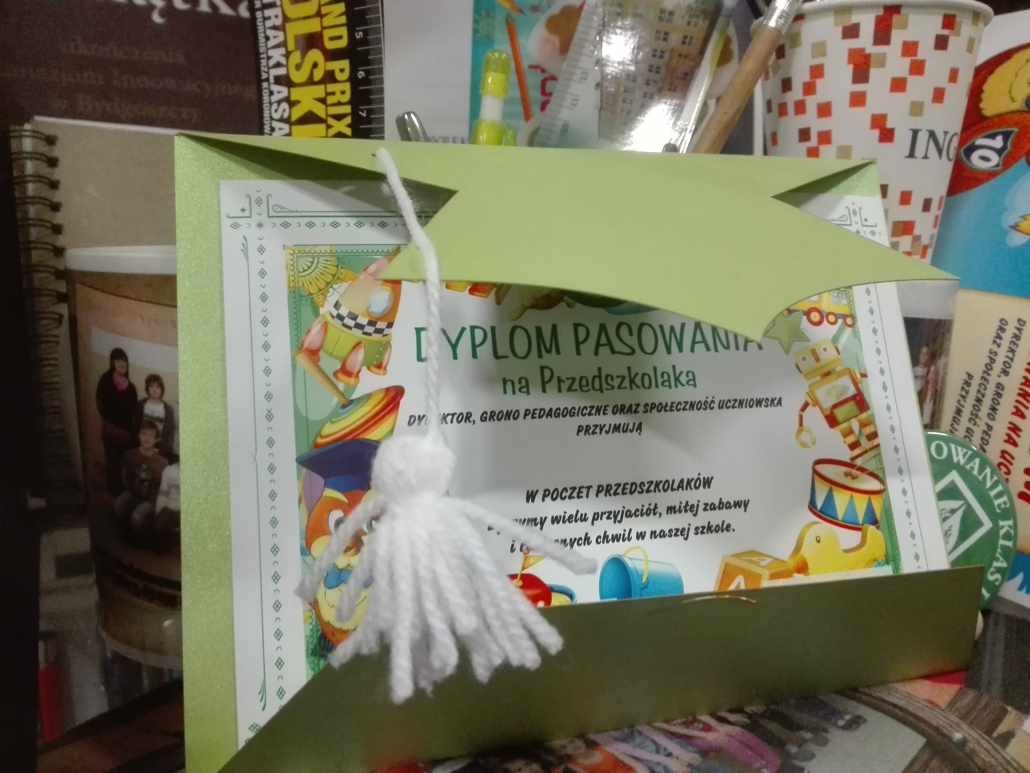 Dyplom pasowania na przedszkolaka z zielonym kartonowym kapeluszem i wełnianym pomponem na tle biurka z różnymi przedmiotami.