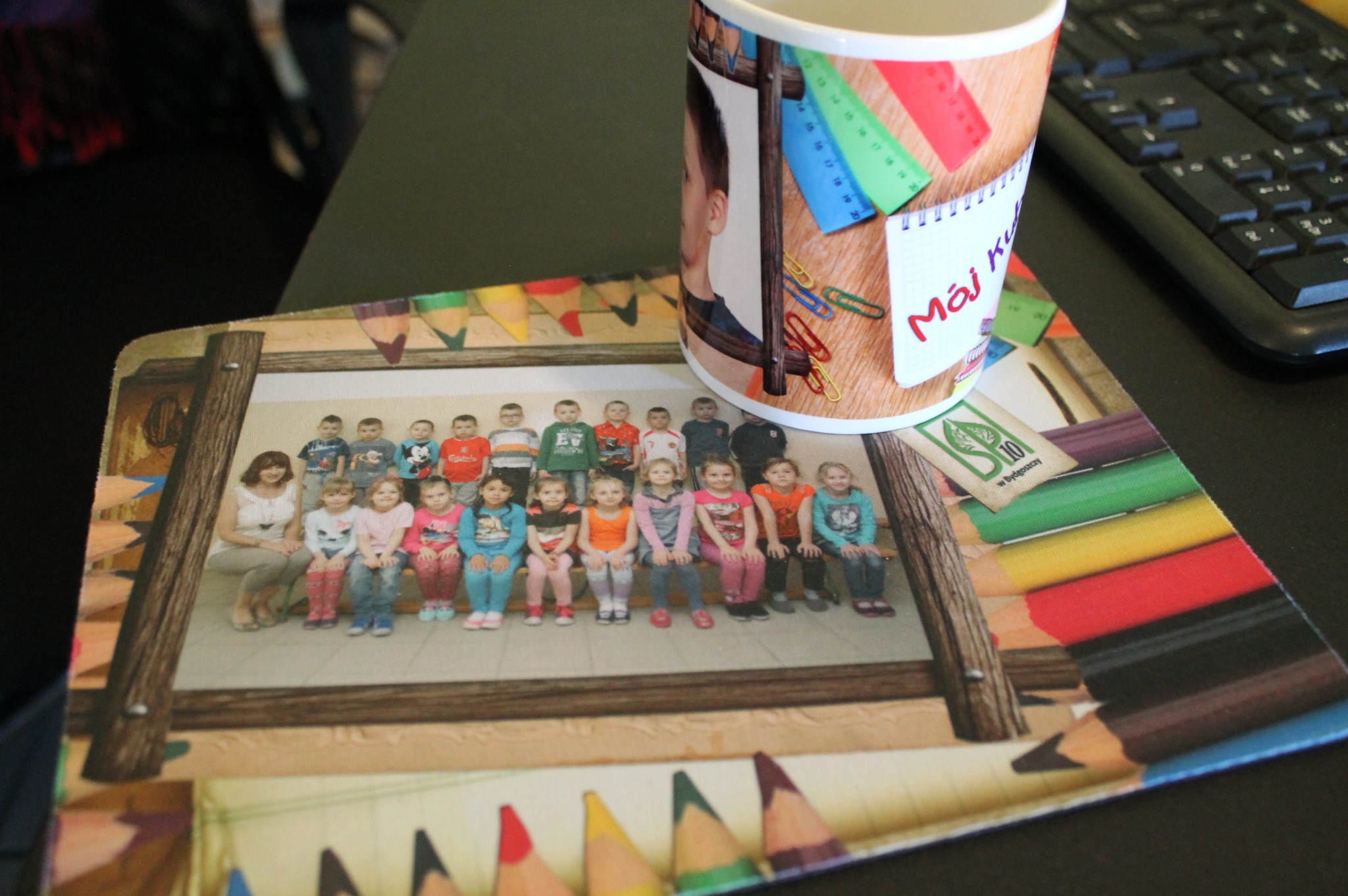 Personalizowany kubek ze zdjęciem chłopca i podkładka pod mysz ze zdjęciem grupowym dzieci i motywem kredek, leżące na biurku obok klawiatury.