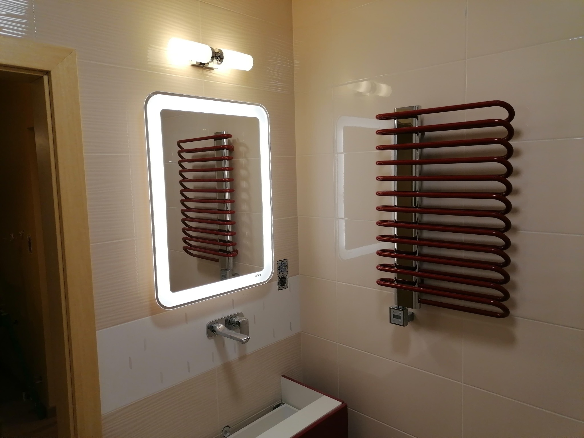 Łazienka po remoncie z podświetlanym lustrem i czerwonym grzejnikiem drabinkowym na tle beżowych płytek, widoczne elementy armatury i fragment drzwi.