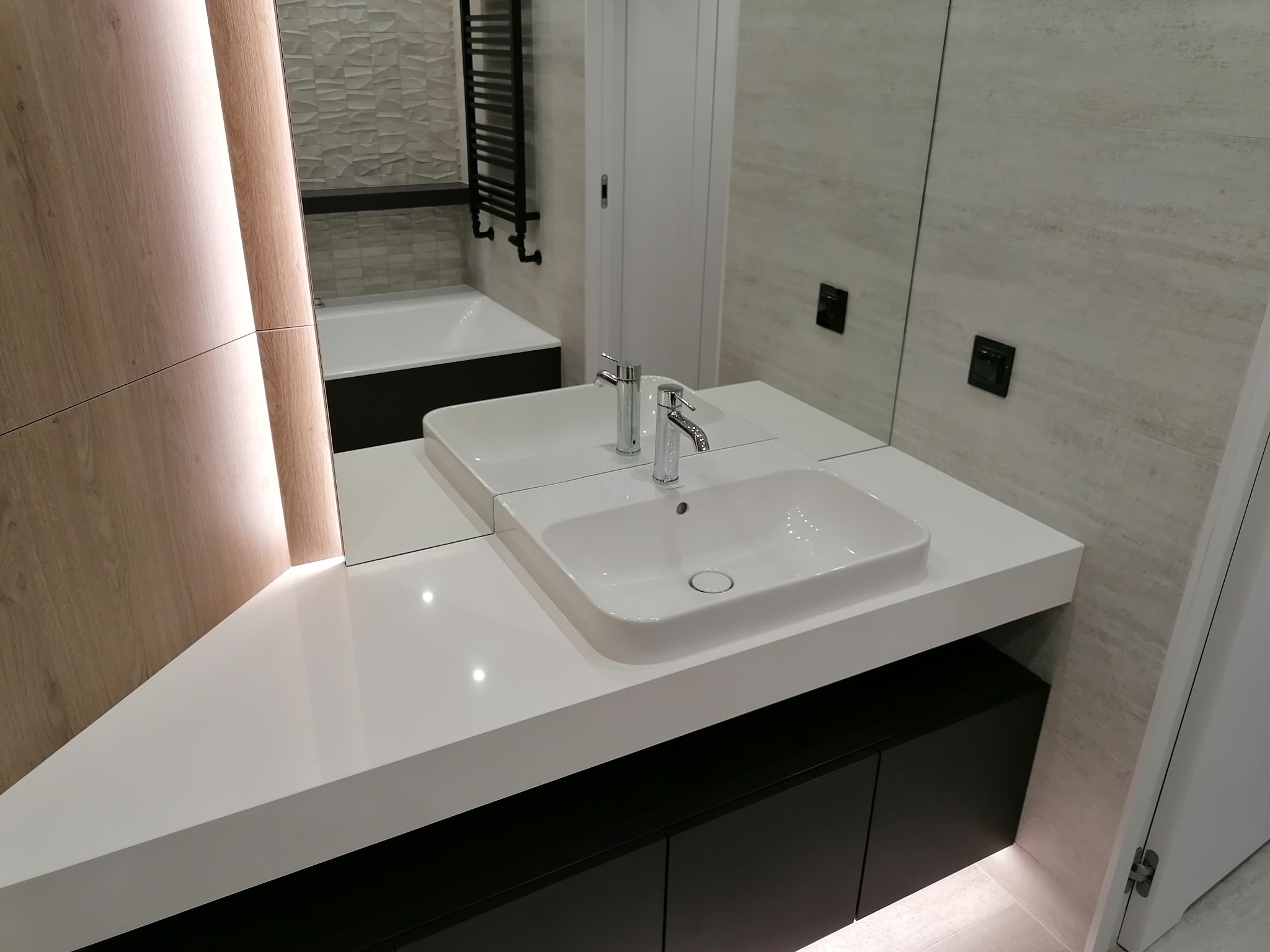 Nowoczesna łazienka z białą umywalką nablatową na czarnym blacie, podświetlona drewniana zabudowa z lustrem, płytki imitujące kamień na ścianach i podświetlany grzejnik drabinkowy w tle.