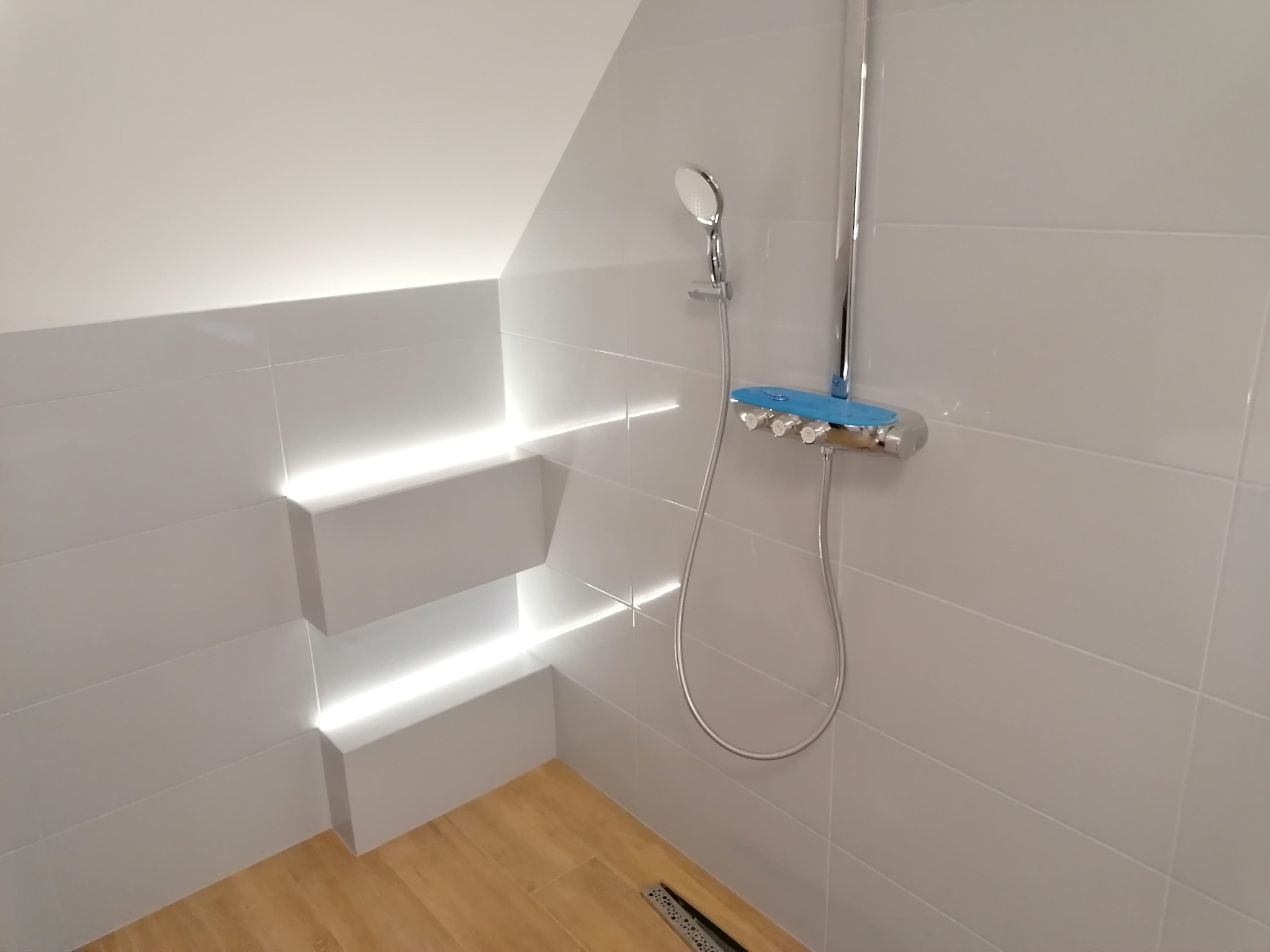 Biała łazienka na poddaszu z nowoczesną armaturą: bateria natryskowa z niebieskimi akcentami i deszczownicą. Ściany wyłożone białymi płytkami z podświetleniem LED w półkach.