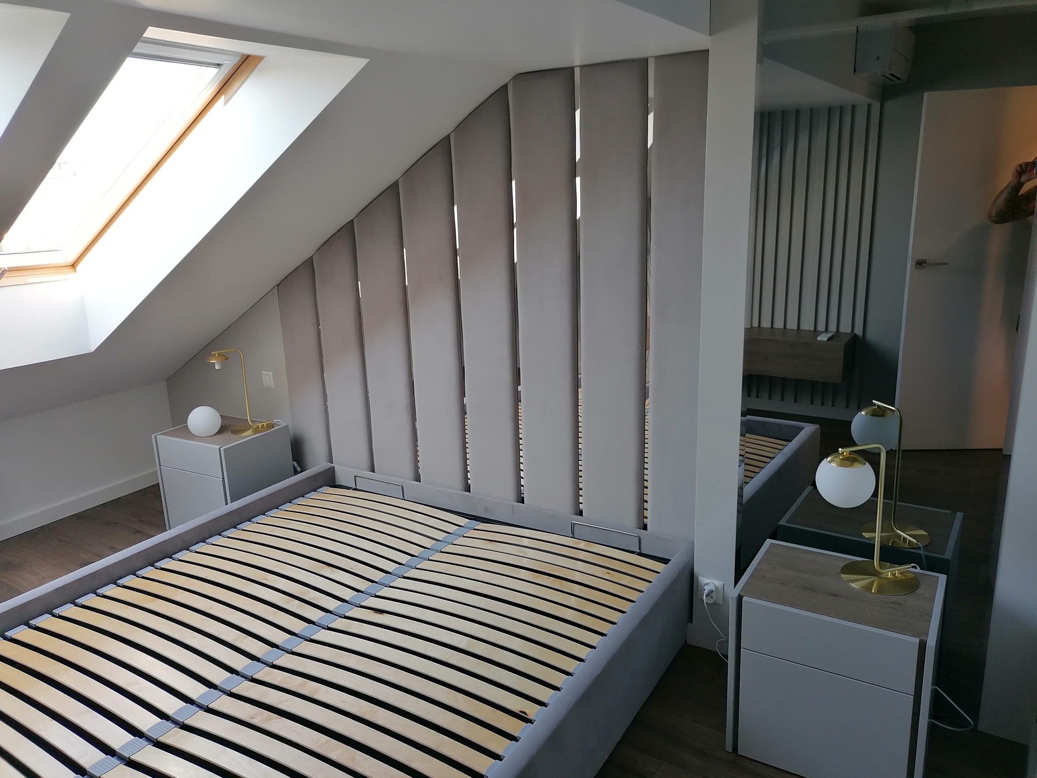 Sypialnia na poddaszu z łóżkiem z szarym, tapicerowanym zagłówkiem, drewnianym stelażem i białymi szafkami nocnymi z lampkami.