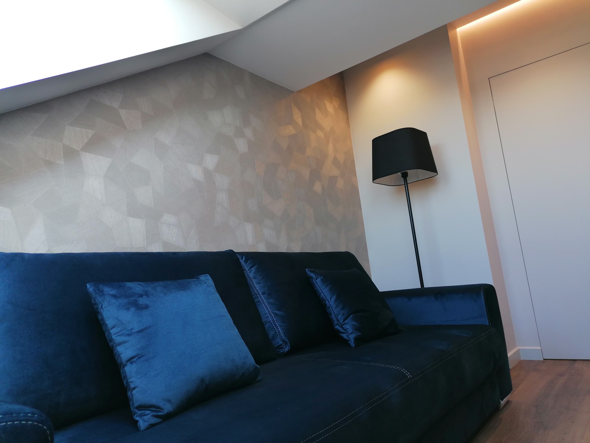 Salon na poddaszu z granatową sofą, dekoracyjną tapetą w geometryczne wzory, lampą podłogową i ukrytym oświetleniem LED wzdłuż ściany.