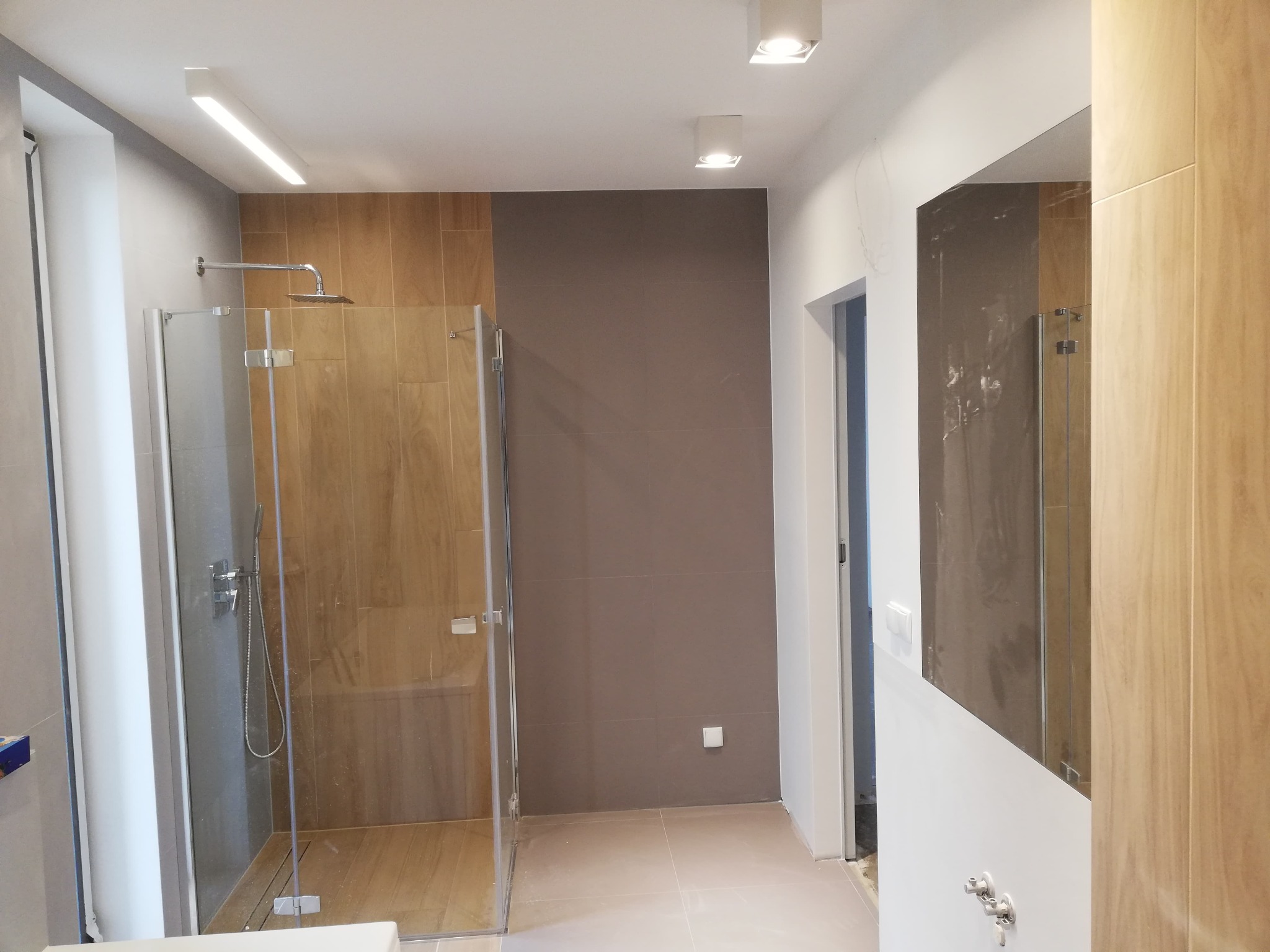 Nowoczesna łazienka z kabiną prysznicową typu walk-in z chromowaną armaturą, wykończona płytkami w odcieniach brązu i beżu, oświetlona punktowymi lampami sufitowymi i listwą LED.