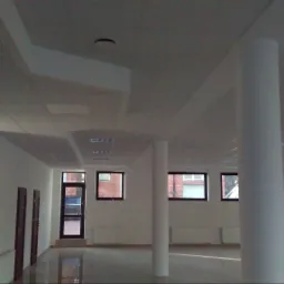 Wnętrze biurowe po remoncie: białe ściany, kolumny, okna z ciemnymi ramami, podwieszany sufit z oświetleniem, drabina przystawna oparta o ścianę.