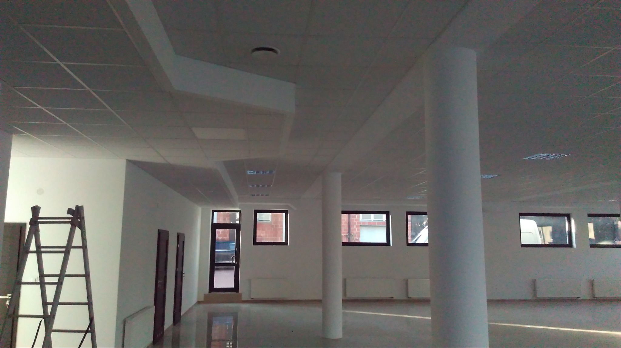 Wnętrze biurowe po remoncie: białe ściany, kolumny, okna z ciemnymi ramami, podwieszany sufit z oświetleniem, drabina przystawna oparta o ścianę.