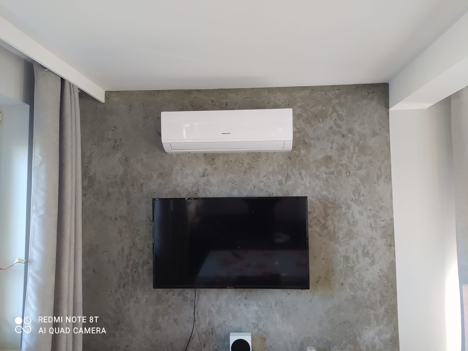 Biały klimatyzator marki Innova zamontowany nad czarnym telewizorem na ścianie o szarym, betonowym wykończeniu w nowoczesnym salonie.