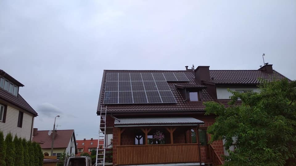 Brązowy dach domu z zainstalowanymi panelami fotowoltaicznymi, drabina oparta o ścianę, pochmurne niebo.
