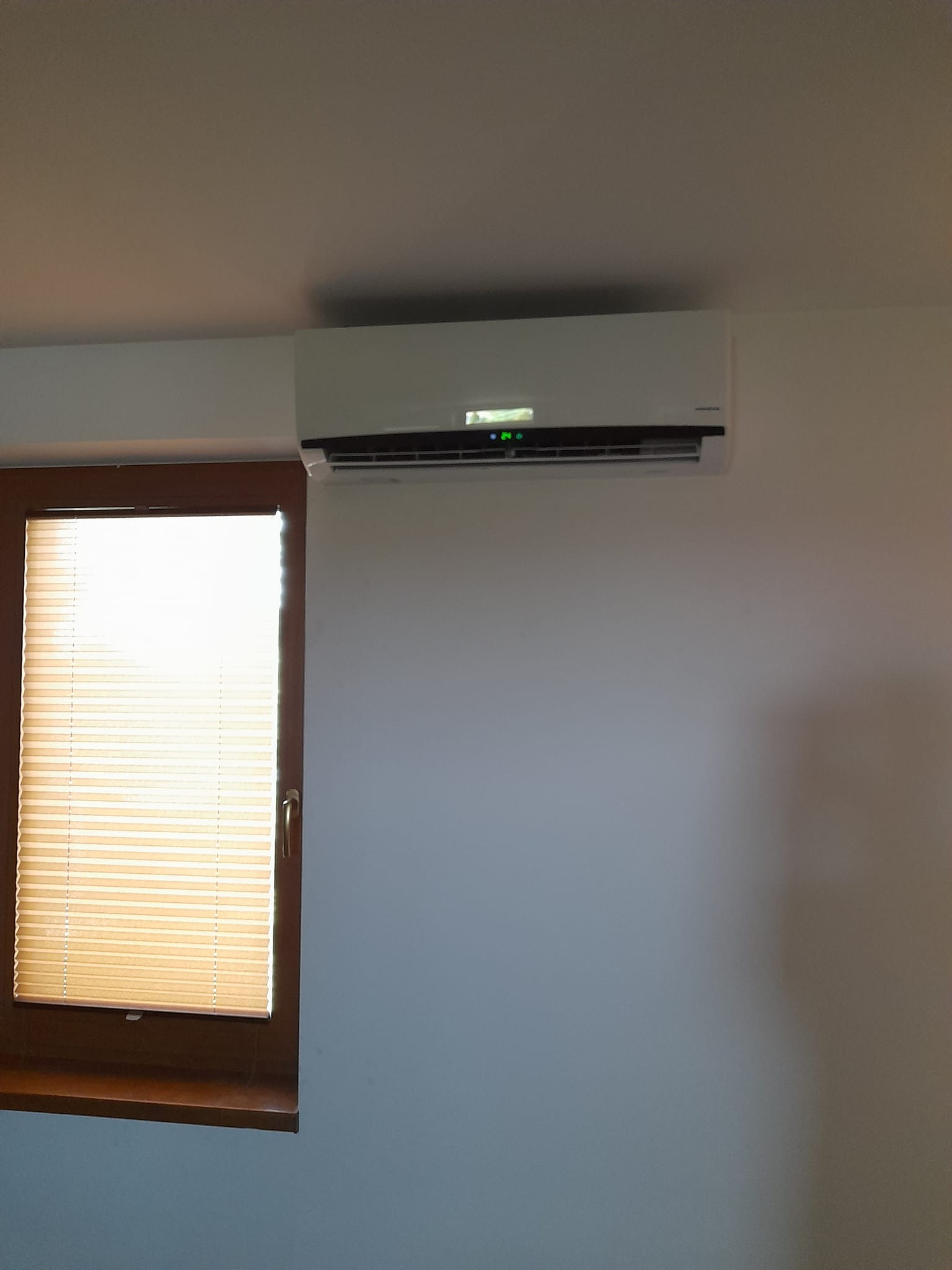 Biały klimatyzator ścienny zamontowany nad oknem z żaluzjami w jasnym pokoju. Na wyświetlaczu klimatyzatora widoczna temperatura 24 stopnie.