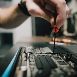 Otwarty laptop z widoczną płytą główną, naprawiany precyzyjnym śrubokrętem z pomarańczową rączką, dłoń serwisanta w górnej części kadru.