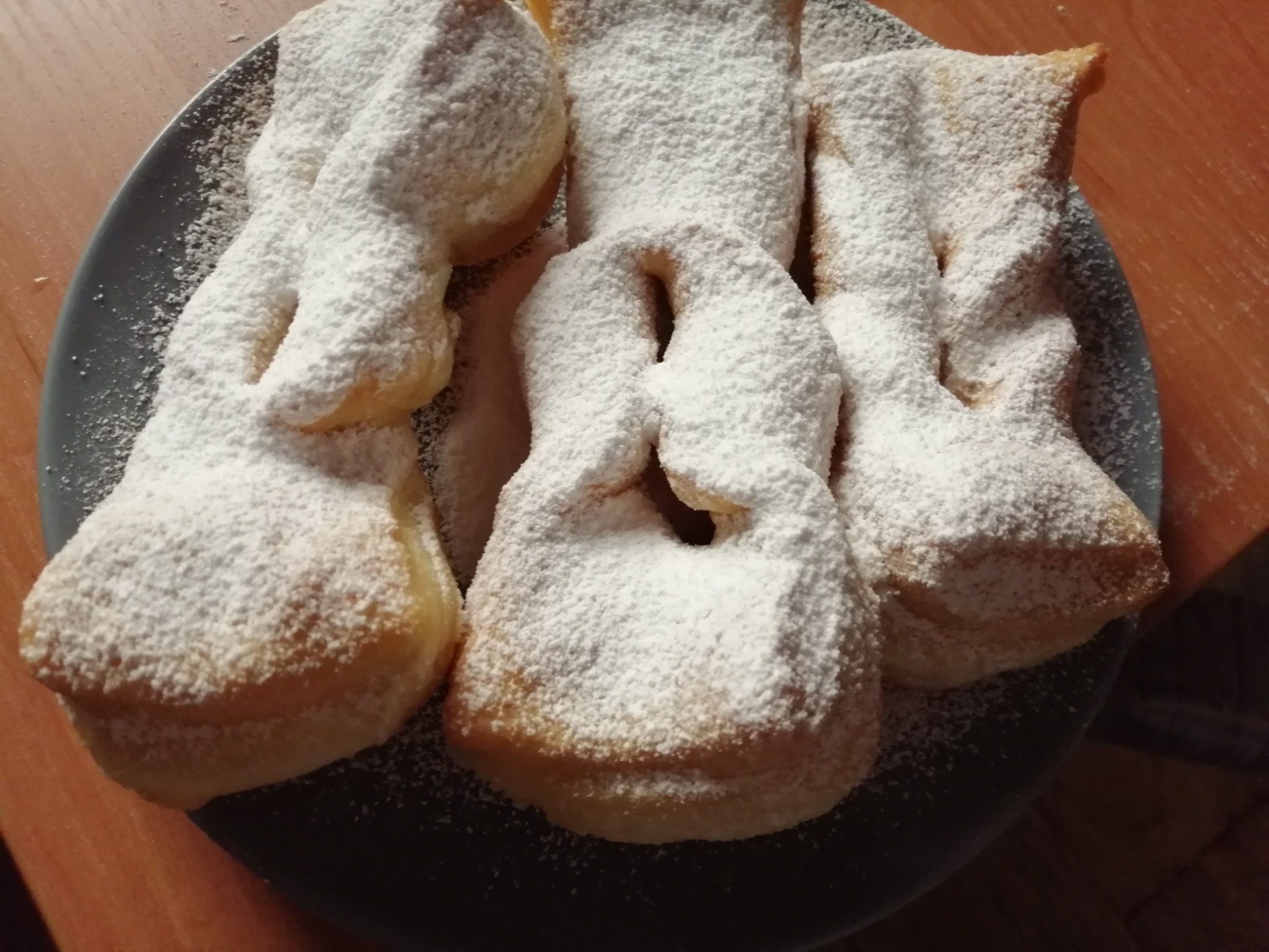 Smażone ciastka faworki, posypane obficie cukrem pudrem, ułożone na szarym talerzu na drewnianym stole.