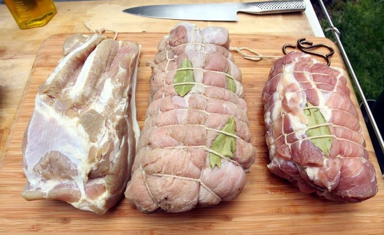 Trzy kawałki surowego mięsa przygotowane do pieczenia lub wędzenia, związane sznurkiem i ozdobione liśćmi laurowymi, leżą na drewnianej desce obok noża.
