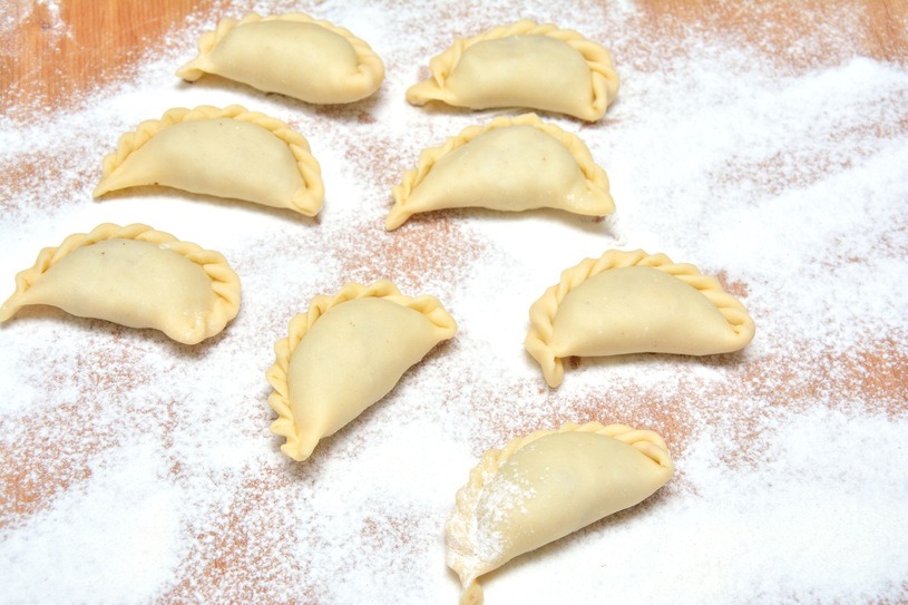 Siedem surowych pierogów w kształcie półksiężyca, z charakterystycznym karbowaniem na brzegach, leżących na drewnianej desce posypanej mąką.