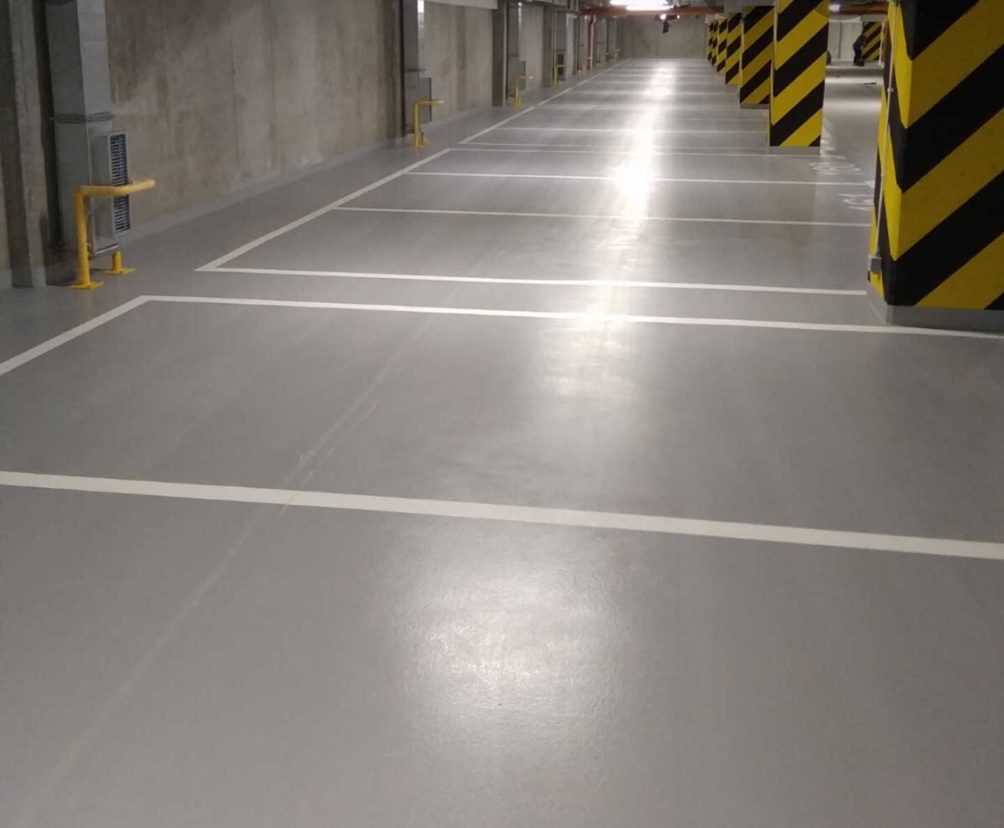 Puste, szare miejsce parkingowe w garażu podziemnym z żółto-czarnymi słupami i białymi liniami wyznaczającymi miejsca postojowe, widok z perspektywy podłogi.