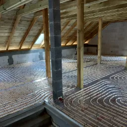 Rozłożone rury instalacji podłogowej na folii izolacyjnej w surowym wnętrzu poddasza, widoczne drewniane krokwie dachu i murowane ściany.