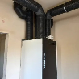 Czarno-biała jednostka Vasco DX6 z podłączonymi czarnymi, karbowanymi rurami wentylacyjnymi zamocowanymi do sufitu, widok w surowym wnętrzu budynku.