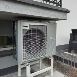 Szara jednostka zewnętrzna z kratką wentylacyjną, zamontowana na białym metalowym stelażu przy ścianie budynku z oknem, obok czarne krzesło z rattanu.