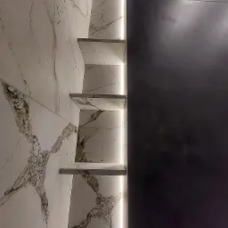 Wykończona ściana łazienki z białych marmurowych płytek z brązowymi żyłami, z wbudowanymi półkami podświetlonymi listwą LED, sąsiadująca z ciemnoszarą, matową ścianą.