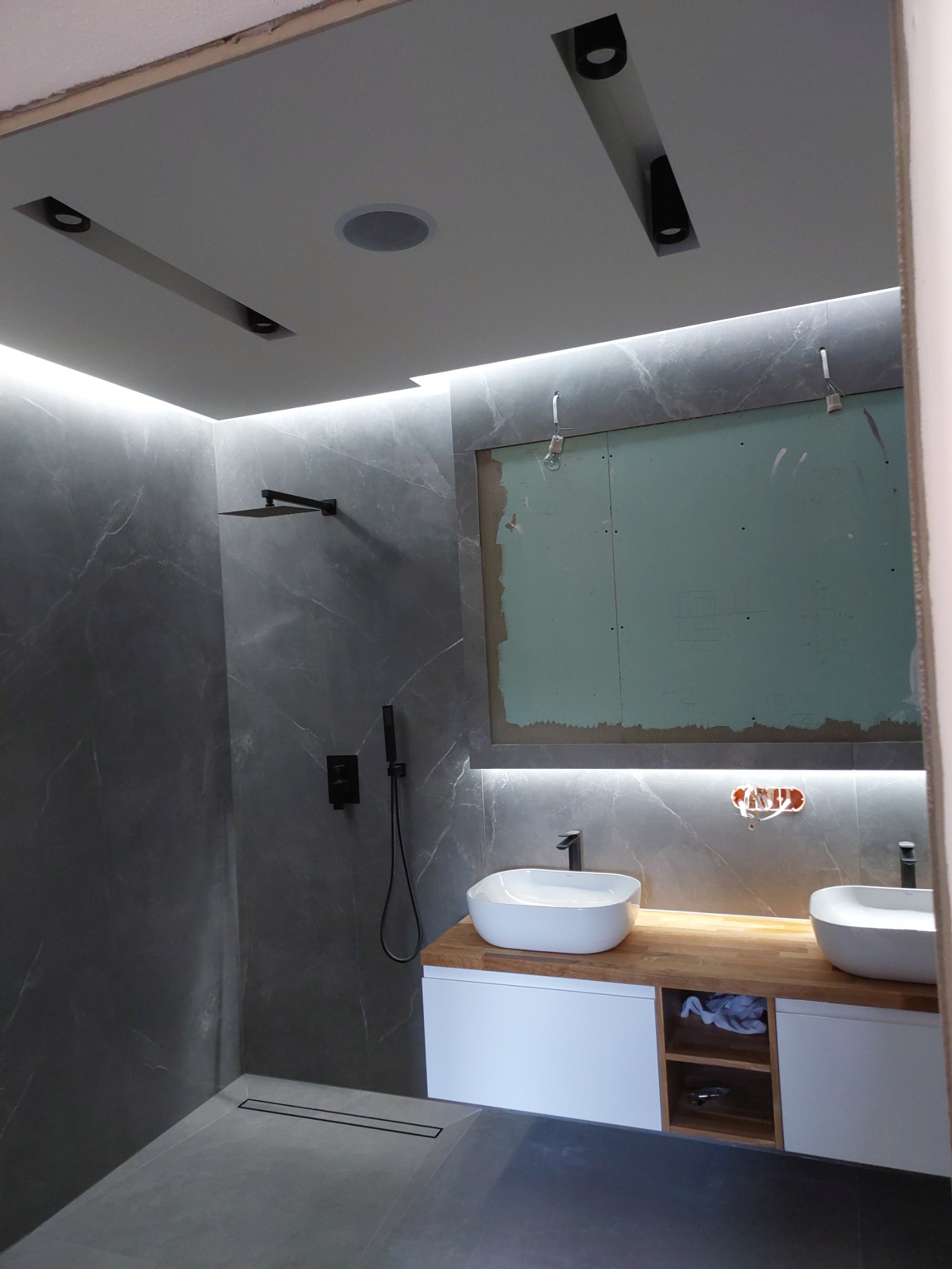 Nowoczesna łazienka w trakcie remontu: szare marmurowe płytki, dwie białe umywalki nablatowe na drewnianym blacie, czarne elementy armatury i oświetlenia, widoczne niedokończone prace przy ścianie.