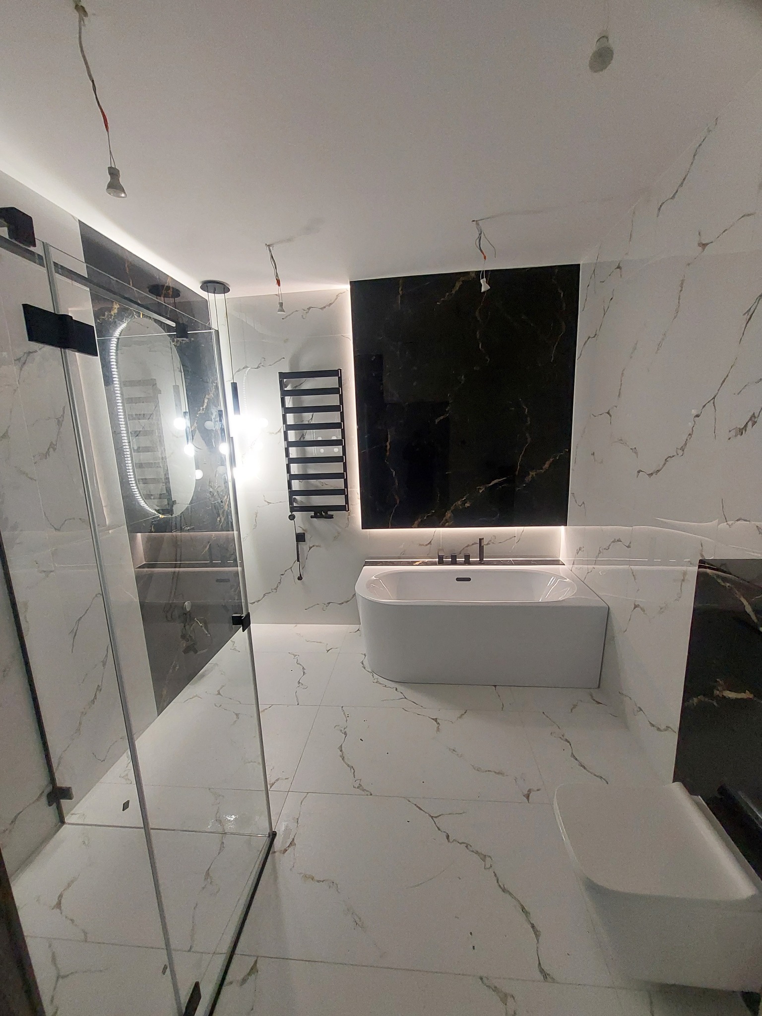 Nowoczesna łazienka z biało-czarnymi marmurkowymi płytkami, kabiną prysznicową typu walk-in, wolnostojącą wanną i grzejnikiem drabinkowym, z widocznymi niedokończonymi instalacjami oświetleniowymi.