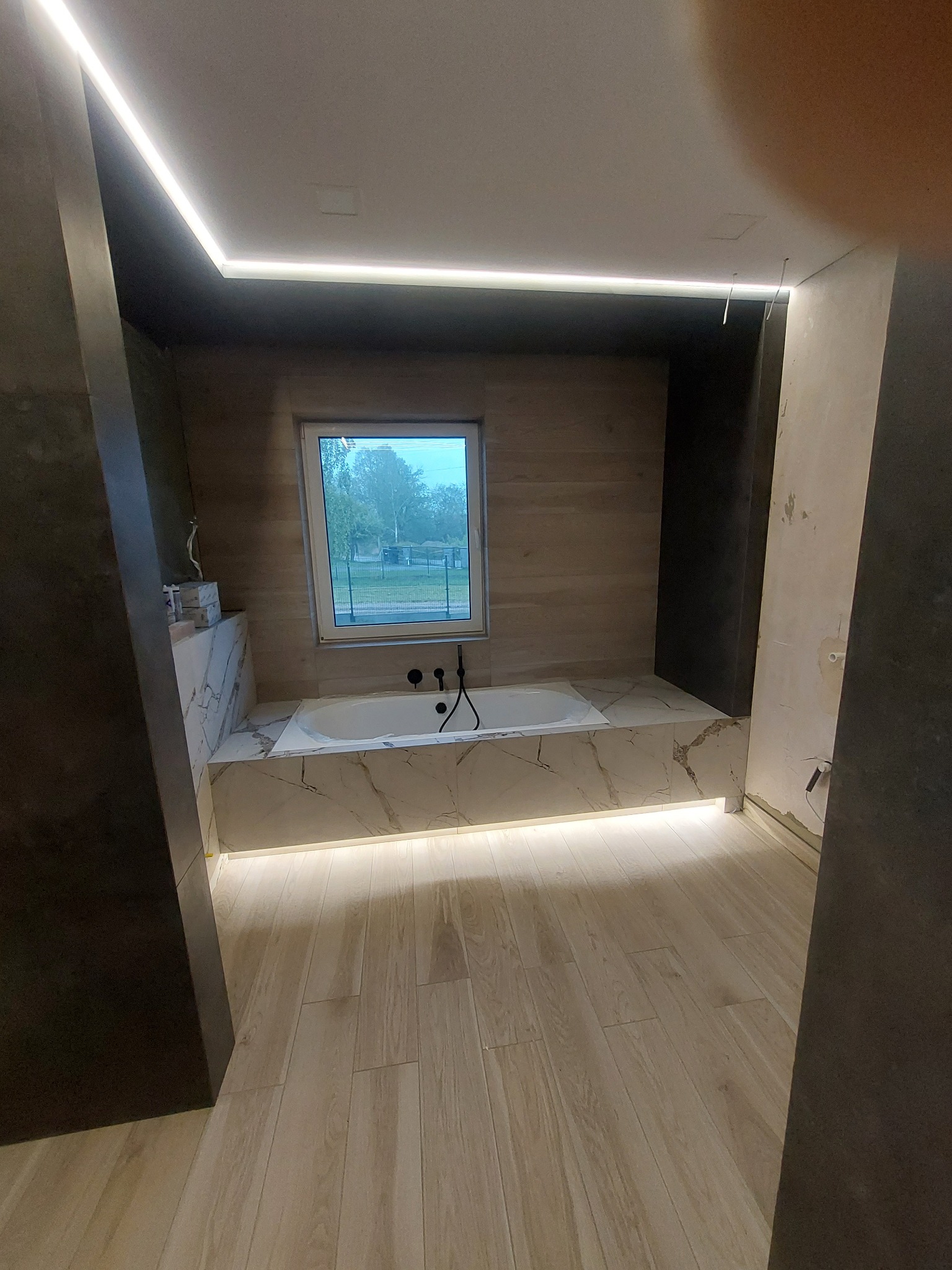Wanna obudowana płytkami imitującymi marmur, z czarną armaturą, oświetlona od dołu i z góry listwami LED, widok na okno i podłogę z paneli imitujących drewno, w trakcie prac wykończeniowych.