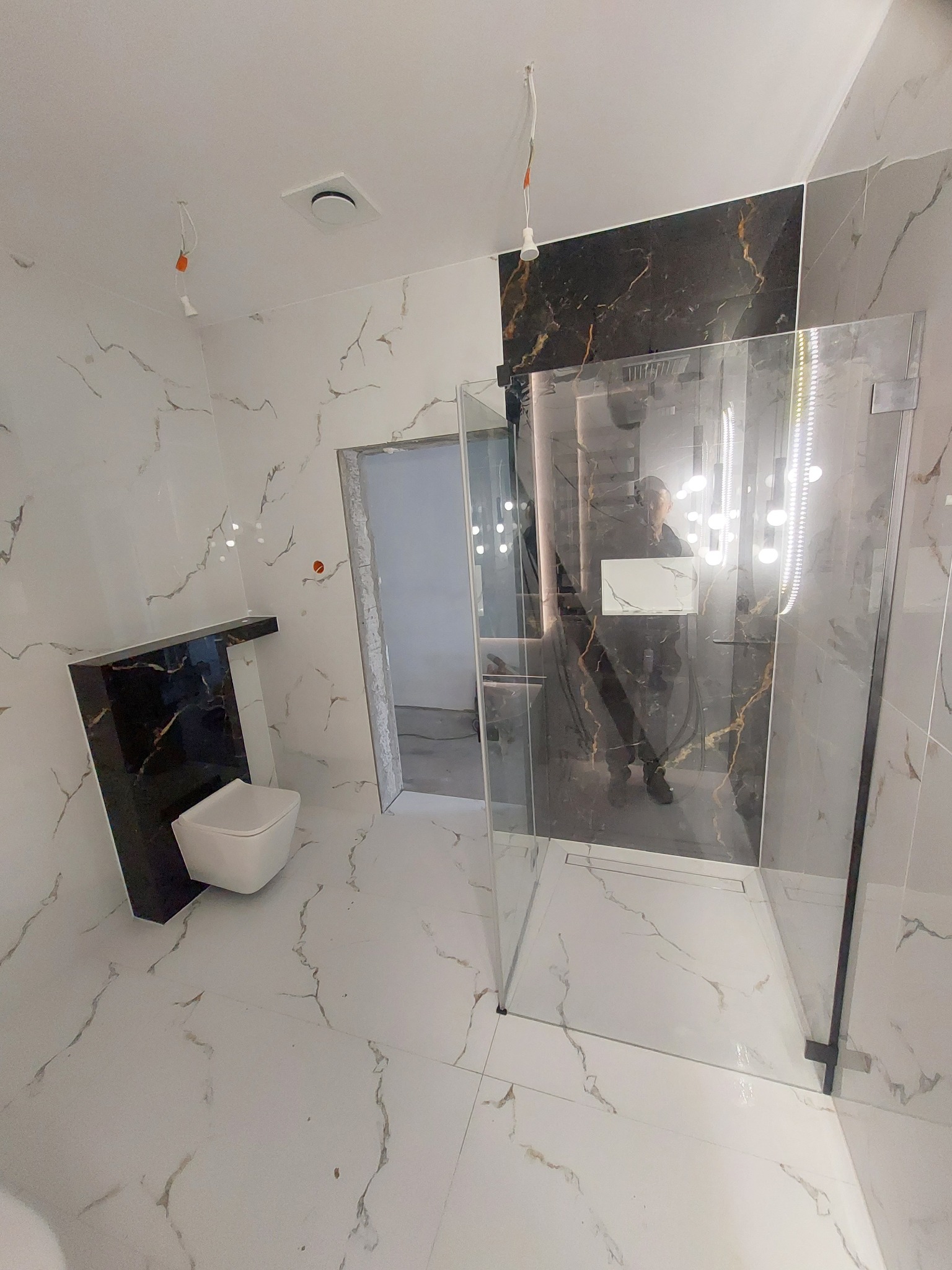 Wykończona łazienka z kabiną prysznicową typu walk-in, białymi płytkami imitującymi marmur z szarymi żyłami, czarnymi akcentami i podwieszaną toaletą.