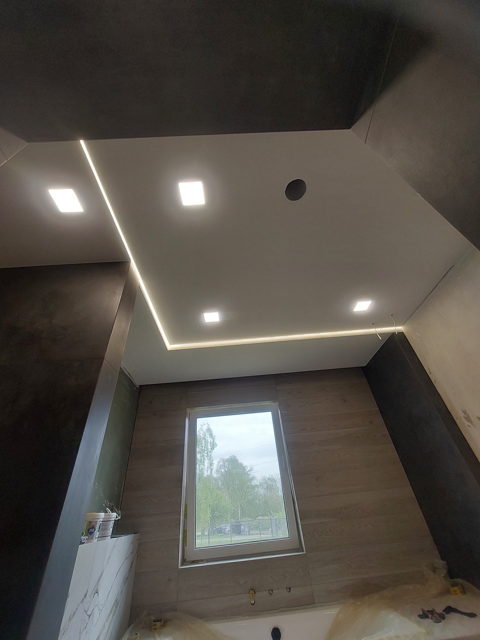 Nowoczesna łazienka w trakcie wykańczania z podwieszanym sufitem z oświetleniem LED, widoczne wiadro z farbą i zabezpieczona folią wanna.