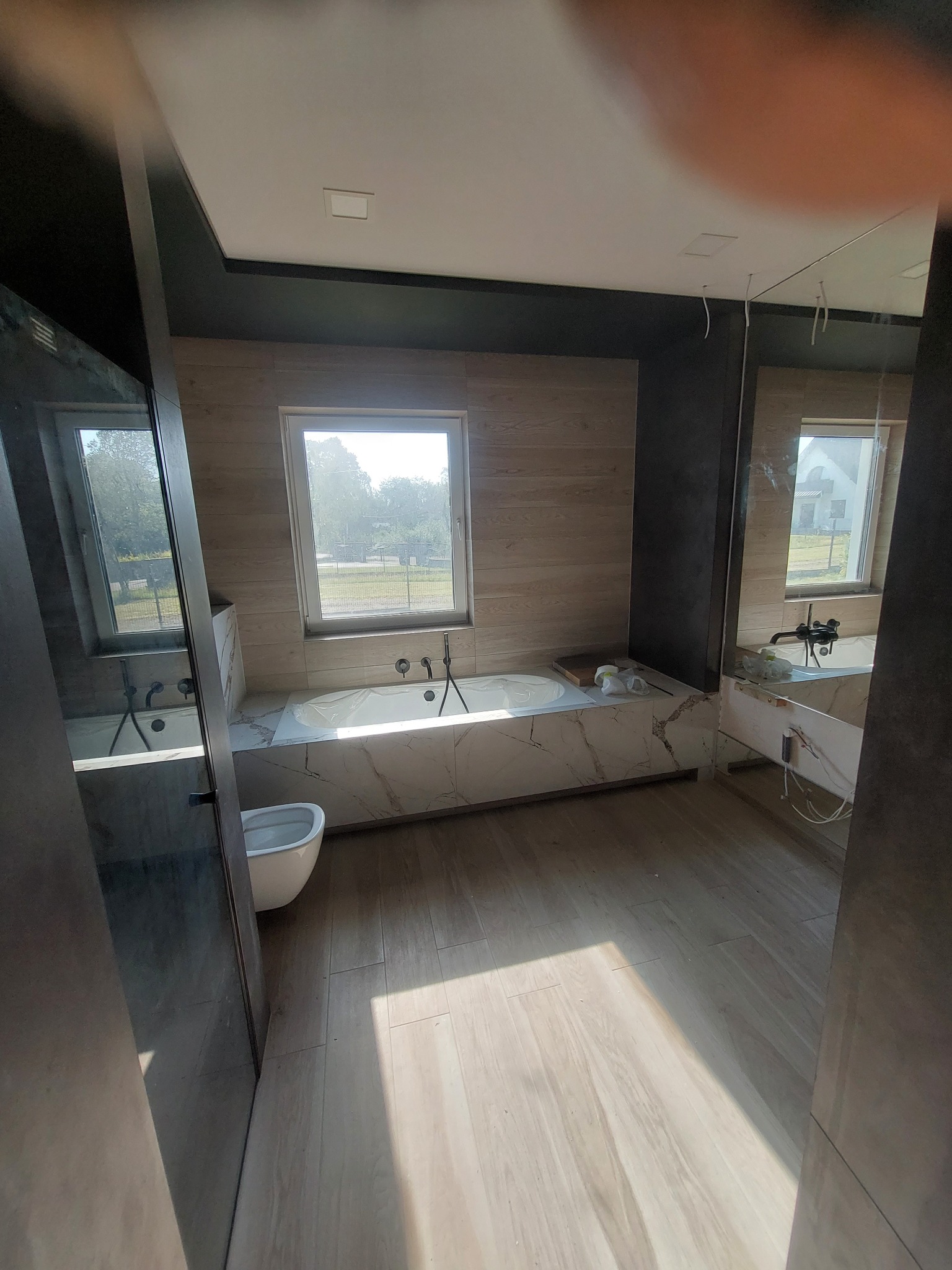 Nowoczesna łazienka z wanną obłożoną marmurem, widok na okno i bidet, drewnopodobne płytki na ścianie, oświetlenie sufitowe oraz lustro z odbiciem fragmentu łazienki.