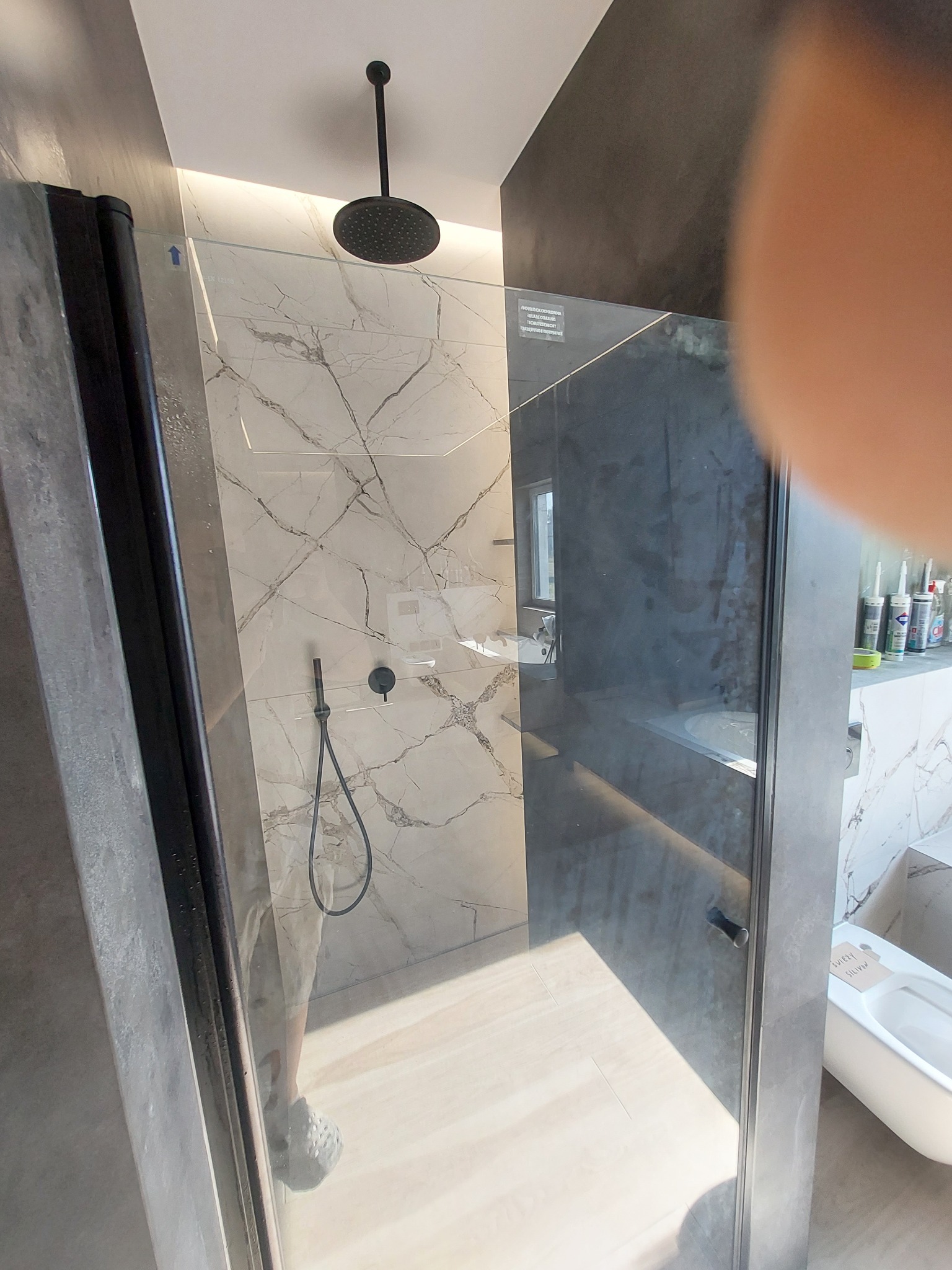Nowoczesna kabina prysznicowa z czarną armaturą, marmurowe płytki na ścianach, widoczna słuchawka prysznicowa i deszczownica, szklane drzwi z czarnymi profilami.
