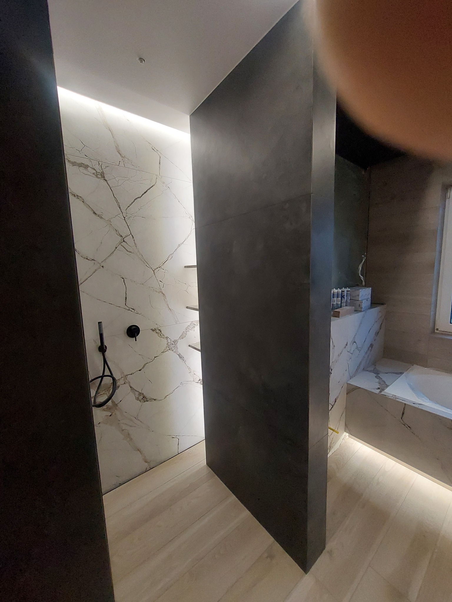 Nowoczesna łazienka z marmurowymi płytkami na ścianach, drewnopodobną podłogą i czarną armaturą, oświetlona ledowym światłem.