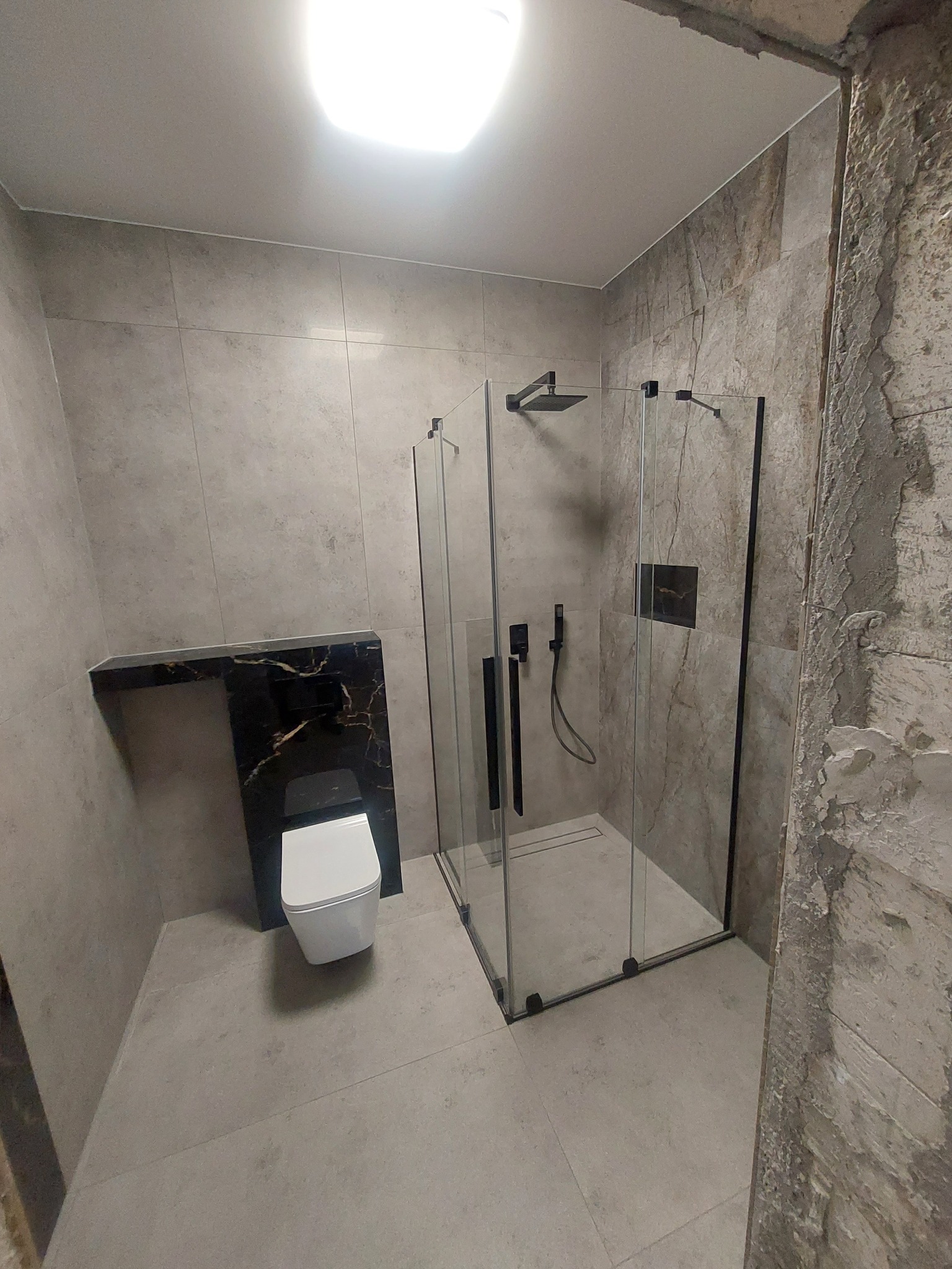 Nowoczesna łazienka w szarościach z kabiną prysznicową typu walk-in z czarnymi okuciami i wiszącą toaletą z czarnym stelażem, widoczne surowe wykończenie narożnika ściany.