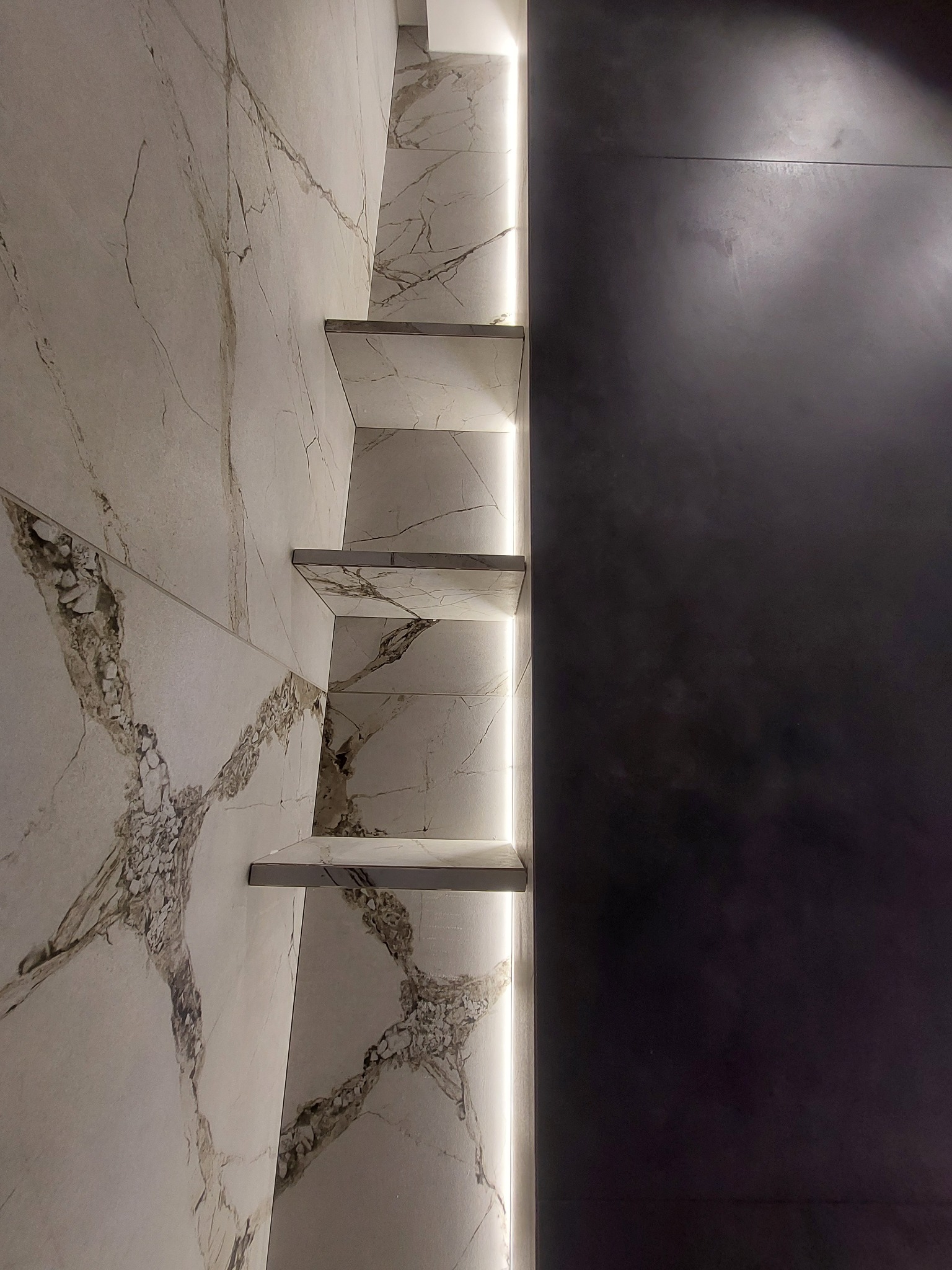 Wykończona ściana łazienki z białych marmurowych płytek z brązowymi żyłami, z wbudowanymi półkami podświetlonymi listwą LED, sąsiadująca z ciemnoszarą, matową ścianą.