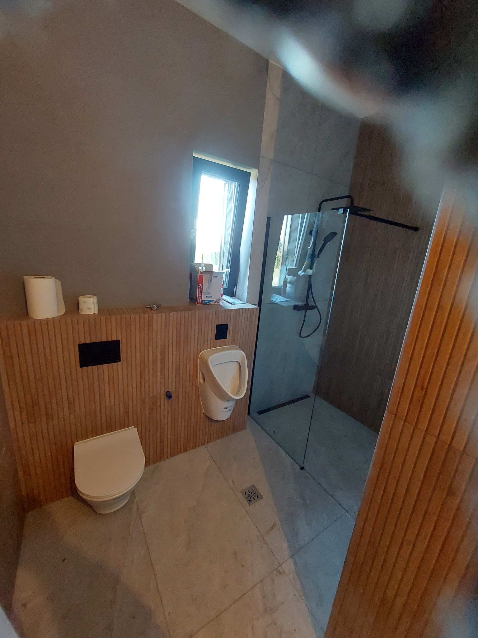 Nowoczesna łazienka z kabiną prysznicową typu walk-in, toaletą i pisuarzem, wykończona płytkami imitującymi drewno na ścianie i szarą podłogą.