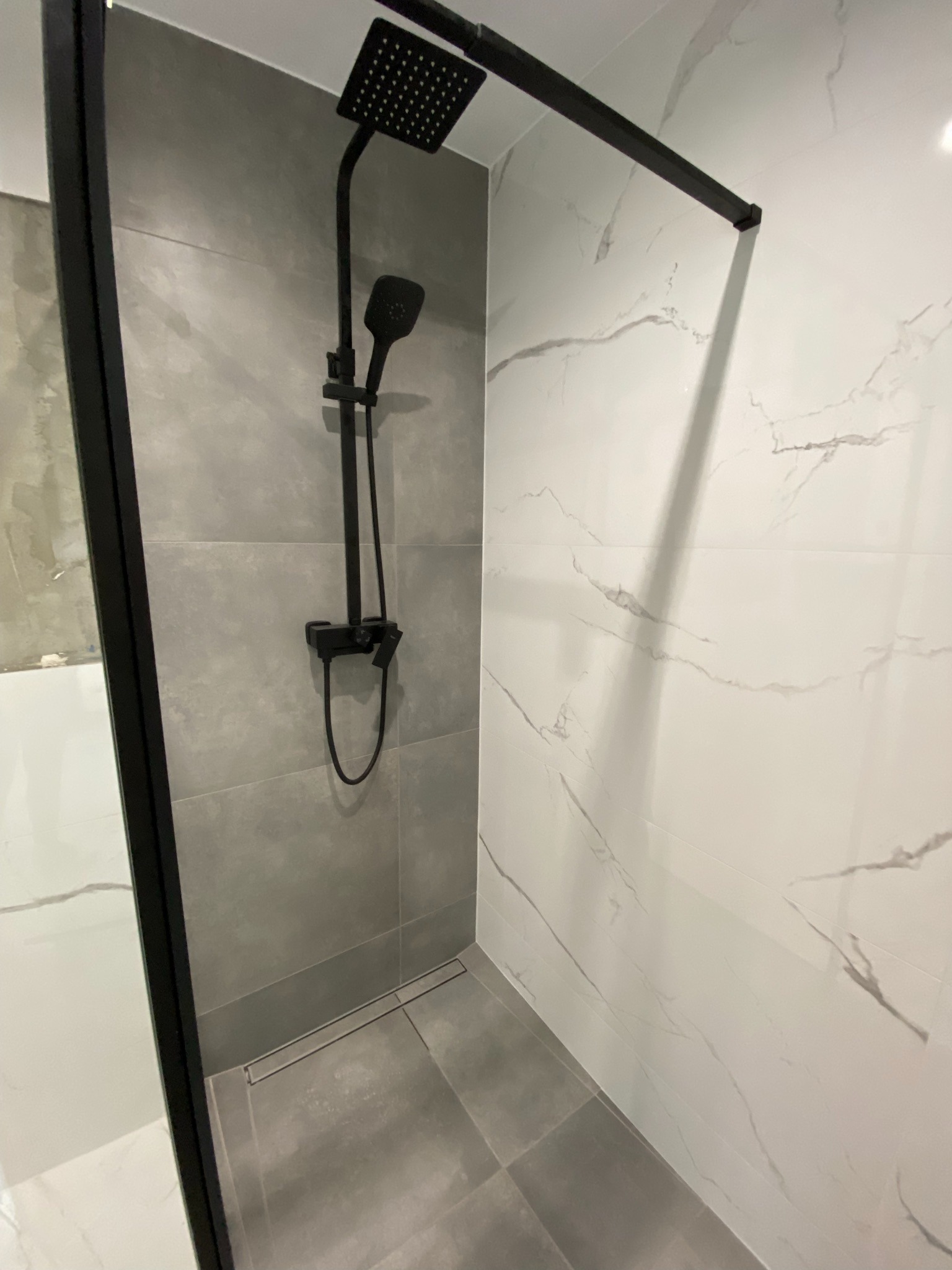 Nowoczesna kabina prysznicowa z czarną armaturą i odpływem liniowym, wykończona płytkami imitującymi marmur i szarym gresem.