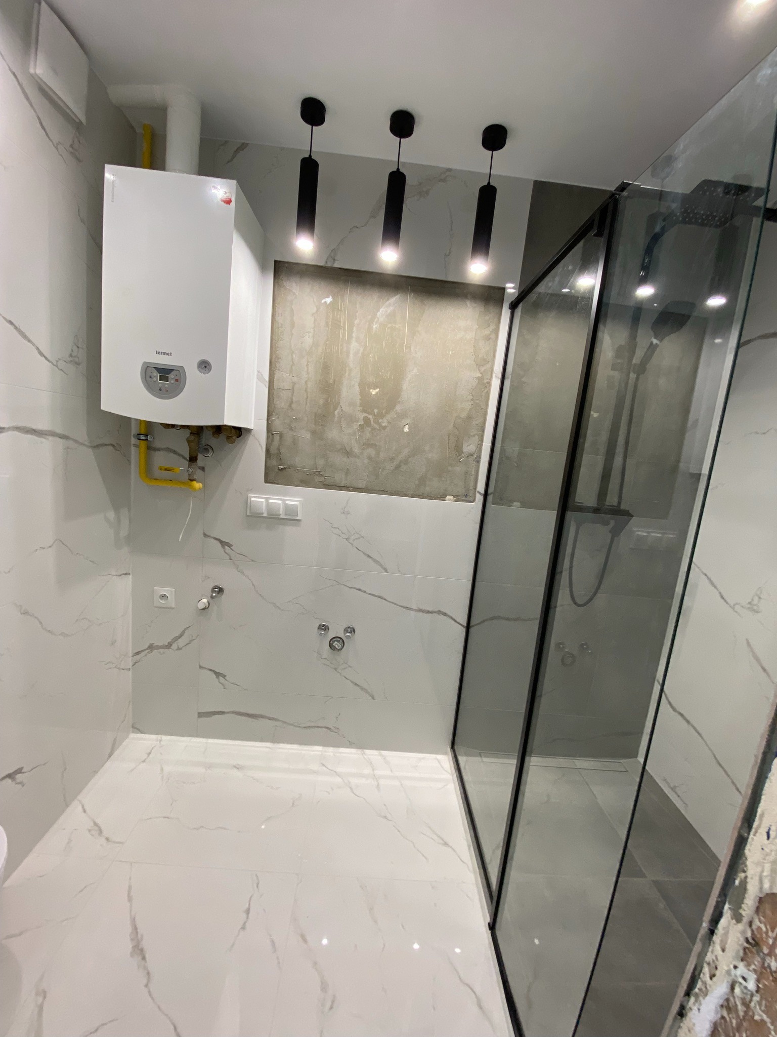 Nowoczesna łazienka z białą marmurową glazurą, kabiną prysznicową typu walk-in z czarnymi ramami, wiszącym piecem gazowym Termet z żółtymi rurami i trzema czarnymi lampami sufitowymi.