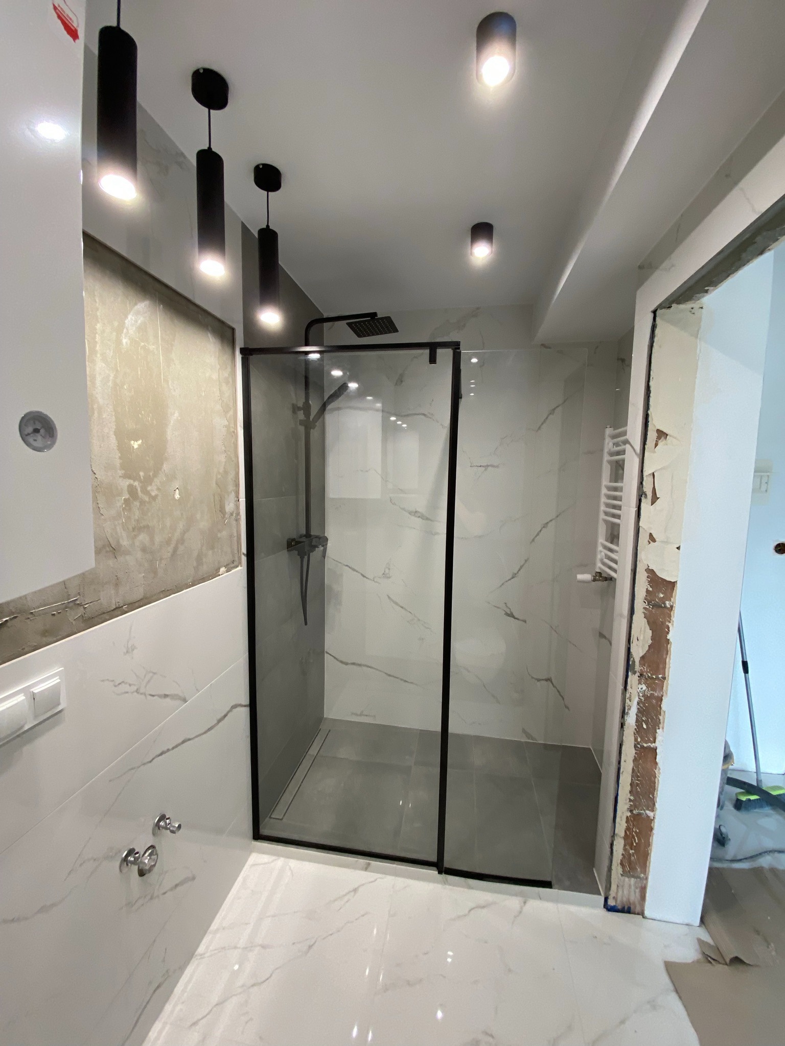 Nowoczesna kabina prysznicowa z czarną ramą i deszczownicą, marmurowe płytki na ścianach i podłodze, czarne lampy sufitowe, widoczny fragment ściany przed położeniem gładzi.