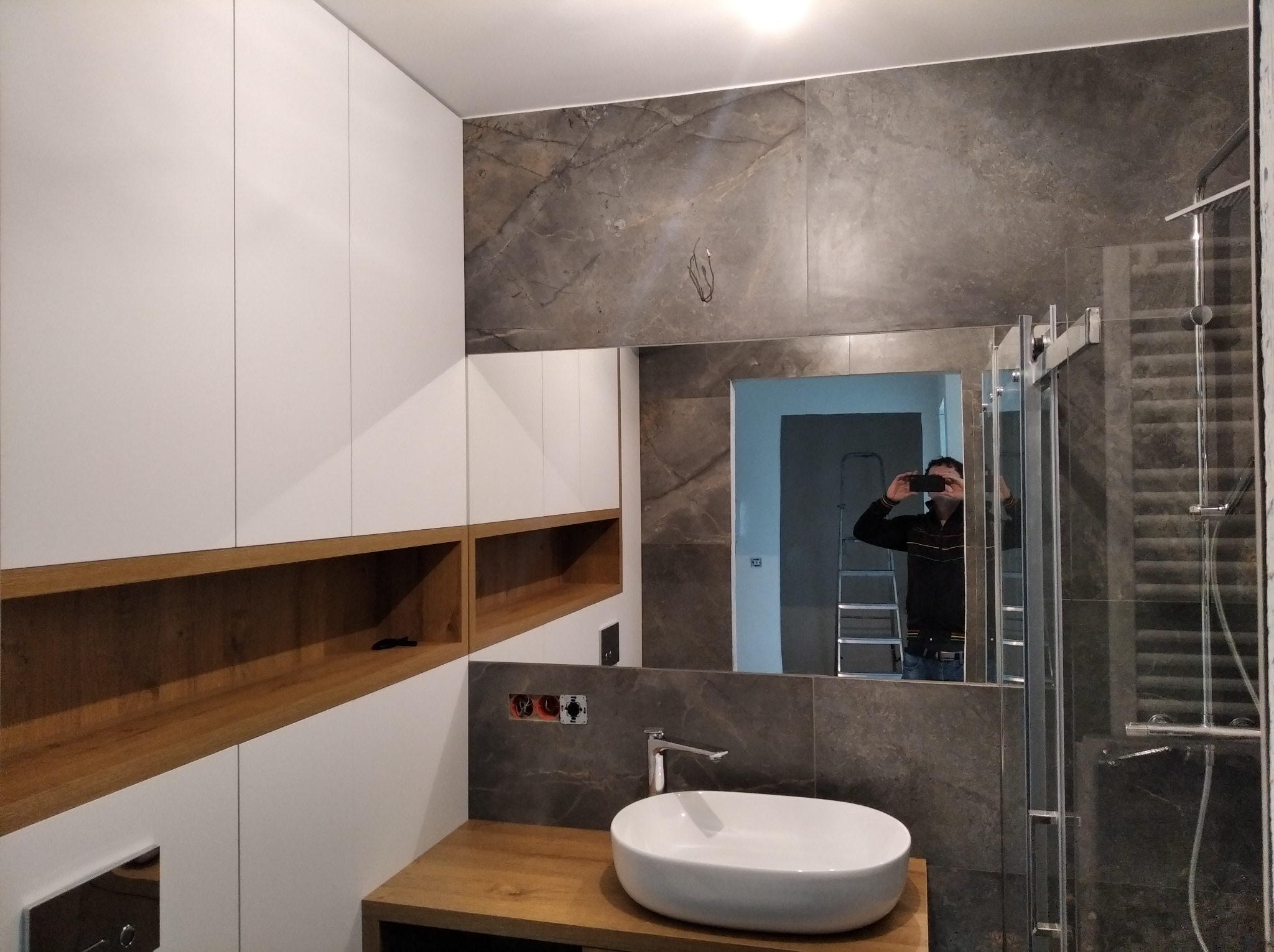 Nowoczesna łazienka z umywalką nablatową, szarą płytką imitującą marmur, białymi szafkami z drewnianymi wnękami i kabiną prysznicową; w lustrze odbija się osoba robiąca zdjęcie i drabina.
