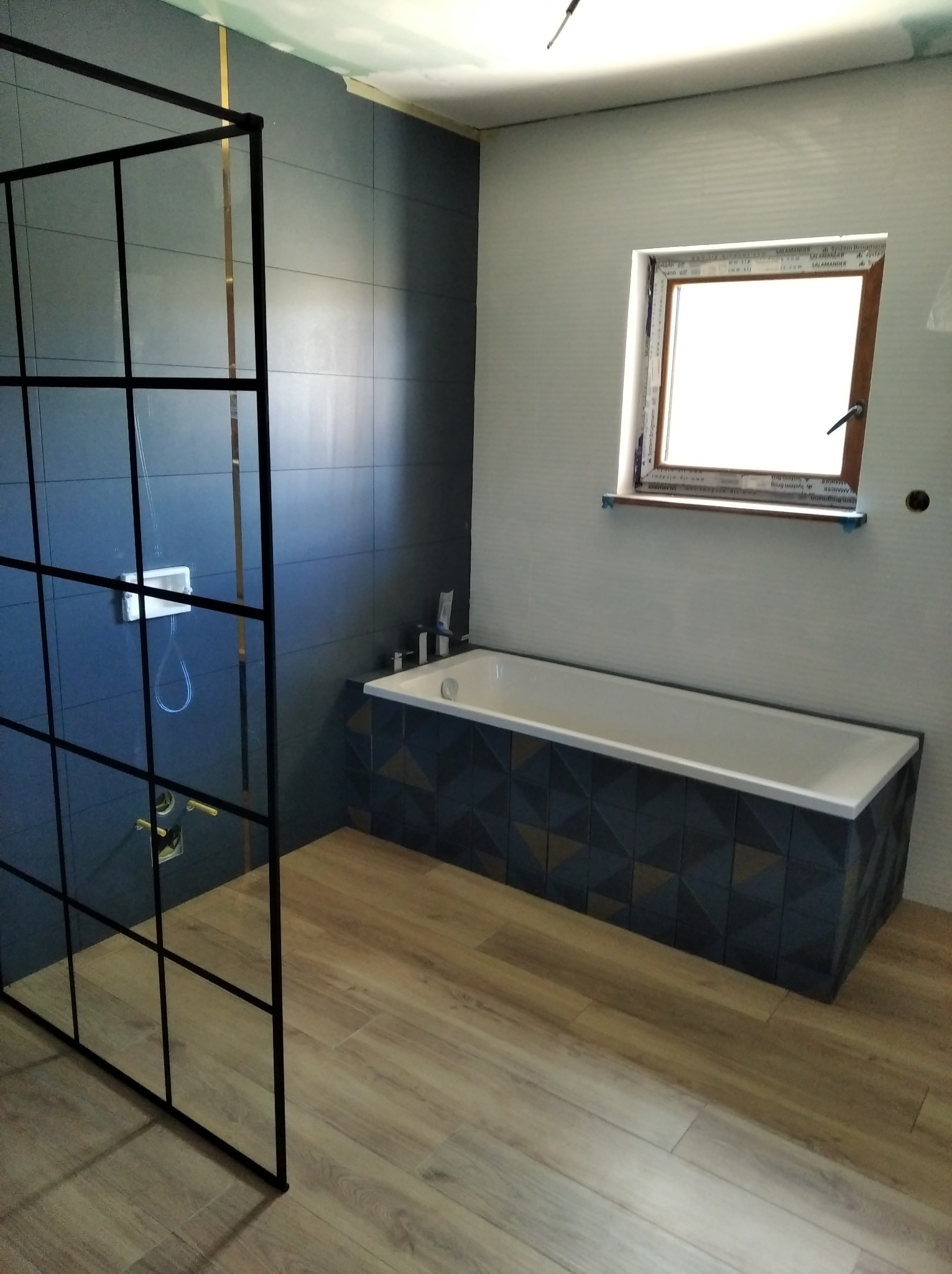 Nowoczesna łazienka w trakcie wykańczania: kabina prysznicowa typu walk-in z czarnymi profilami, ciemne płytki na ścianach z elementami złota, obudowa wanny z geometrycznym wzorem, drewnopodobna...