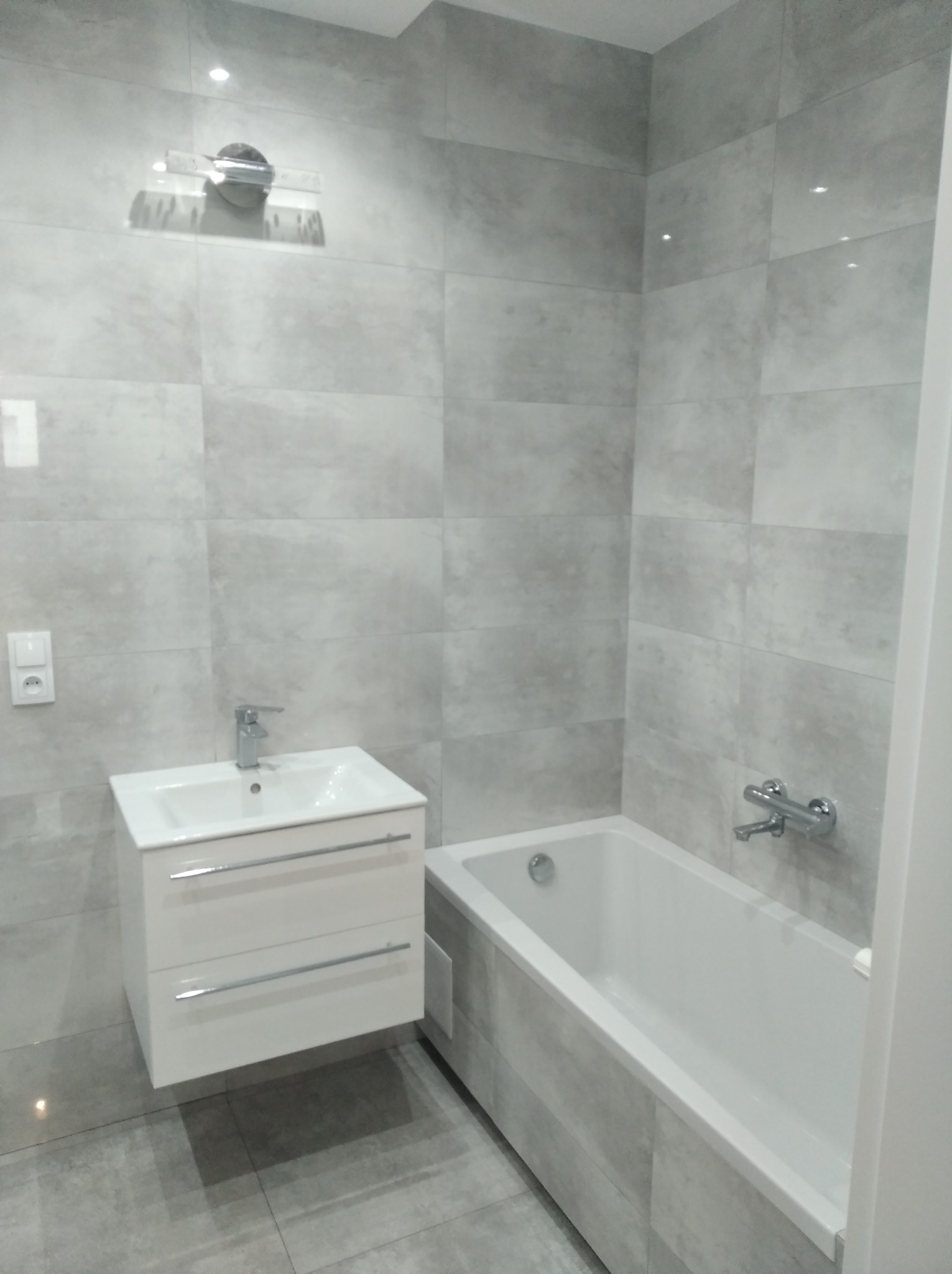 Nowoczesna łazienka z wanną obudowaną szarymi płytkami, białą umywalką z szafką podwieszaną i szarą płytką na ścianach.